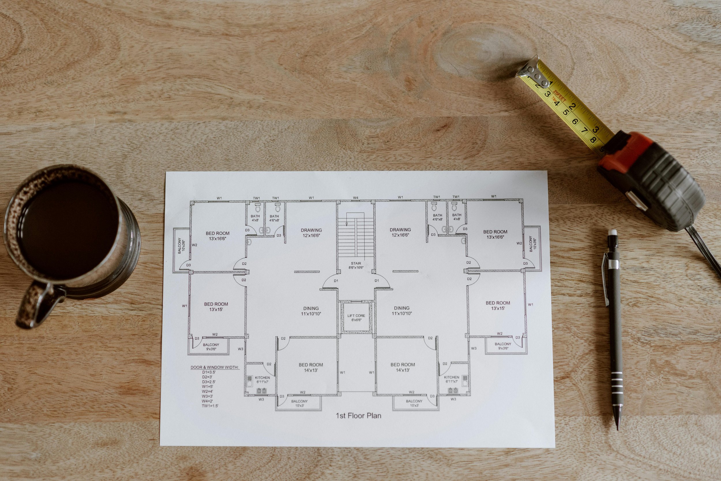 Blueprints of a house floor plan on a wooden table with a tape measure, a pen, and a cup of coffee.
