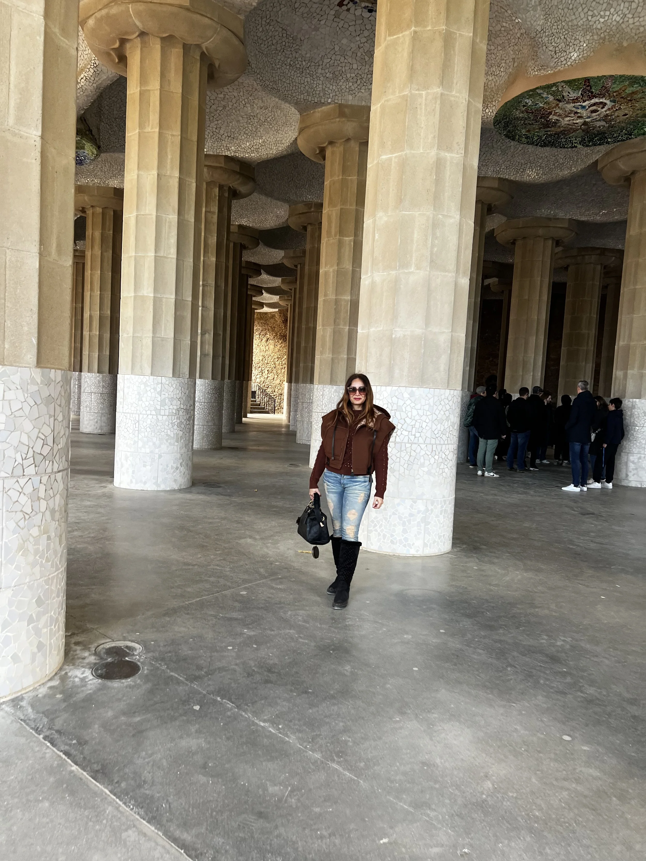 Woman standing inside a large, modern architectural structure with tall, textured stone columns and a mosaic ceiling. She is wearing a brown jacket, ripped jeans, and black boots, holding a black bag, and wearing sunglasses. There are other people in