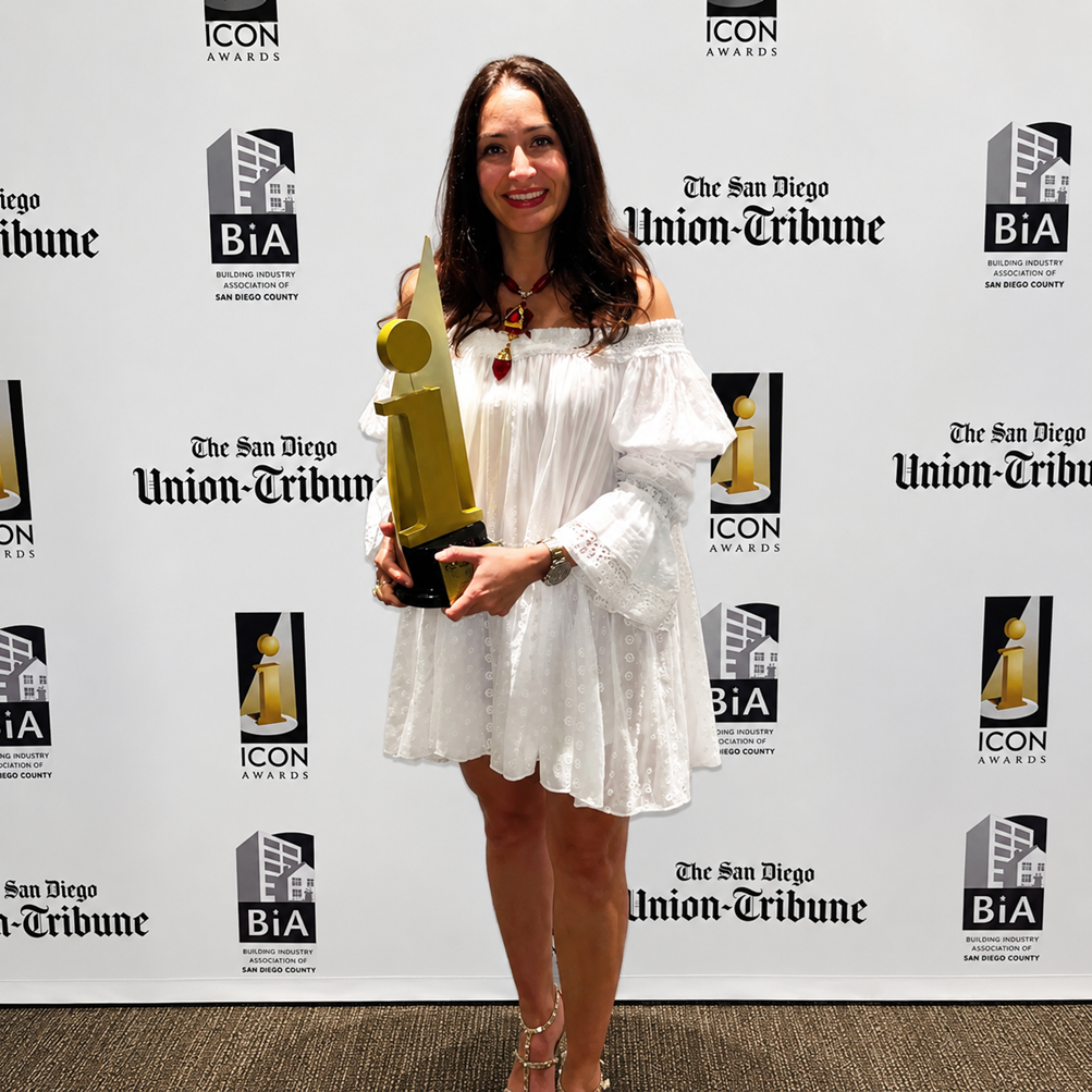 A woman in a white off-shoulder dress standing indoors in front of a backdrop with logos, holding a gold award trophy shaped like the letter 'i'.