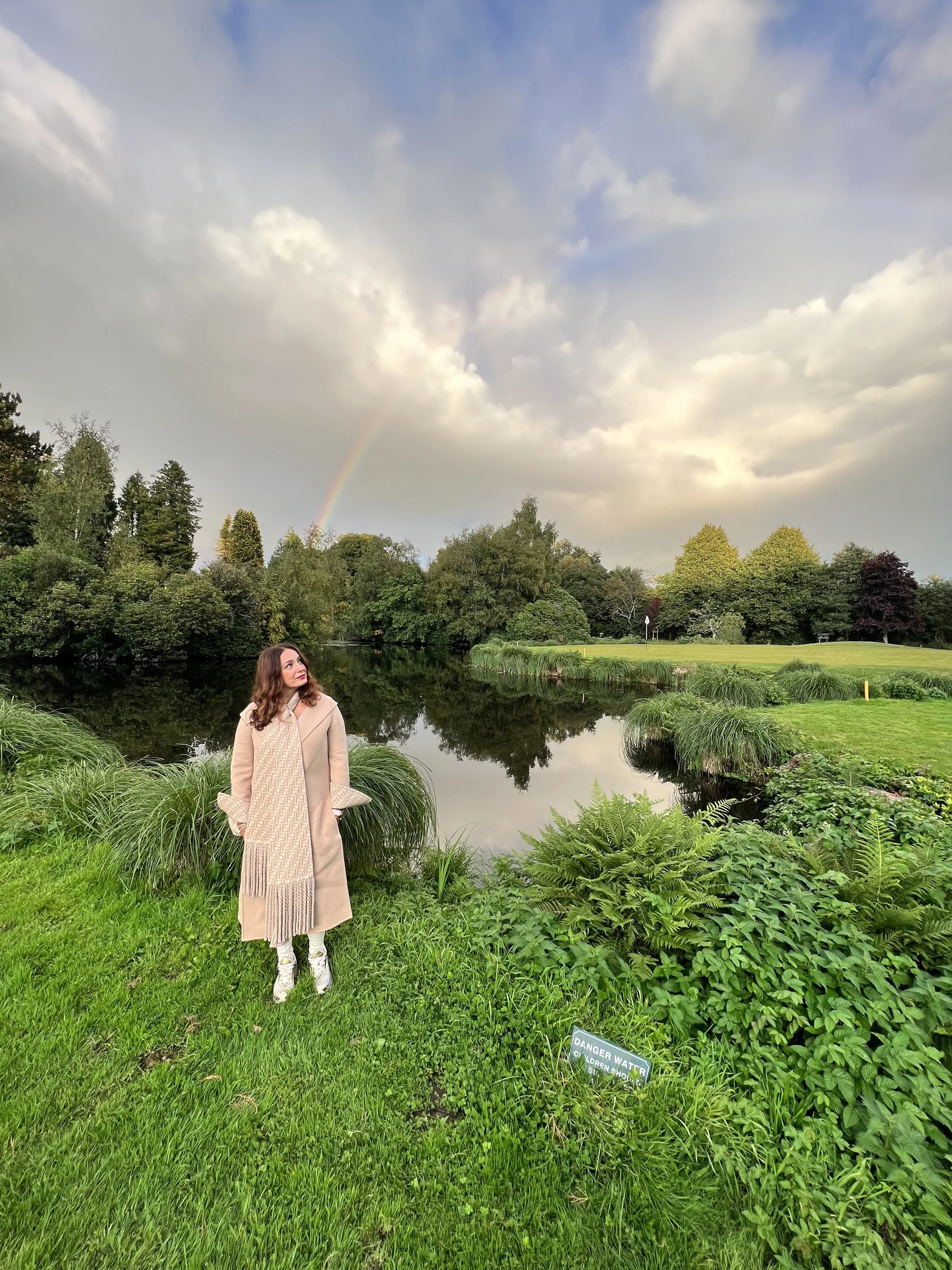 A woman standing near a pond in a lush green park, with a rainbow and cloudy sky overhead.