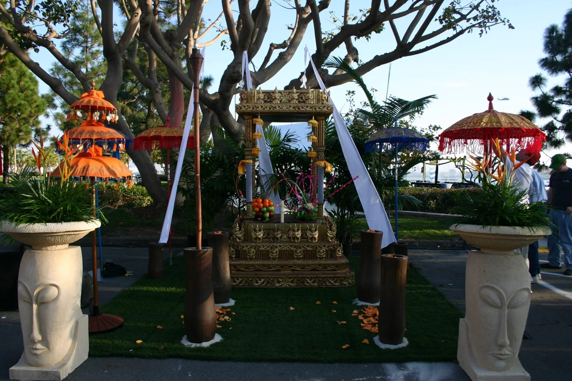 A decorated outdoor altar with an ornate gold structure in the center, surrounded by colorful umbrellas and large planters with plants and flowers, with people and trees in the background.