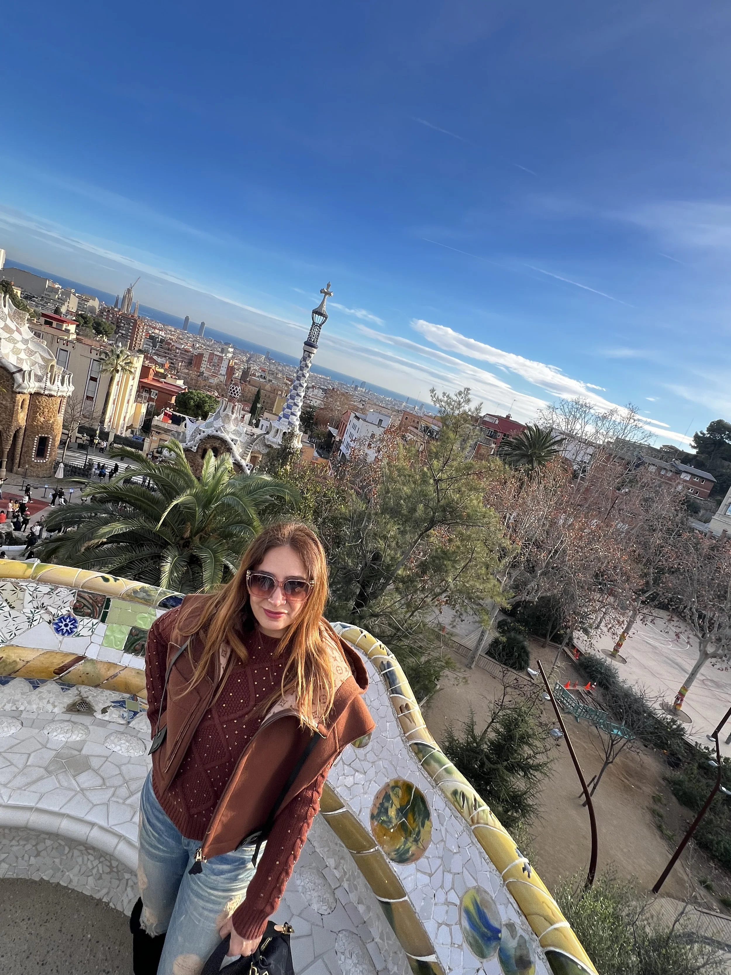 A woman with red hair, wearing sunglasses, a brown jacket, and a maroon sweater, standing on a mosaic bench in Park Güell, Barcelona, with the cityscape and blue sky in the background.