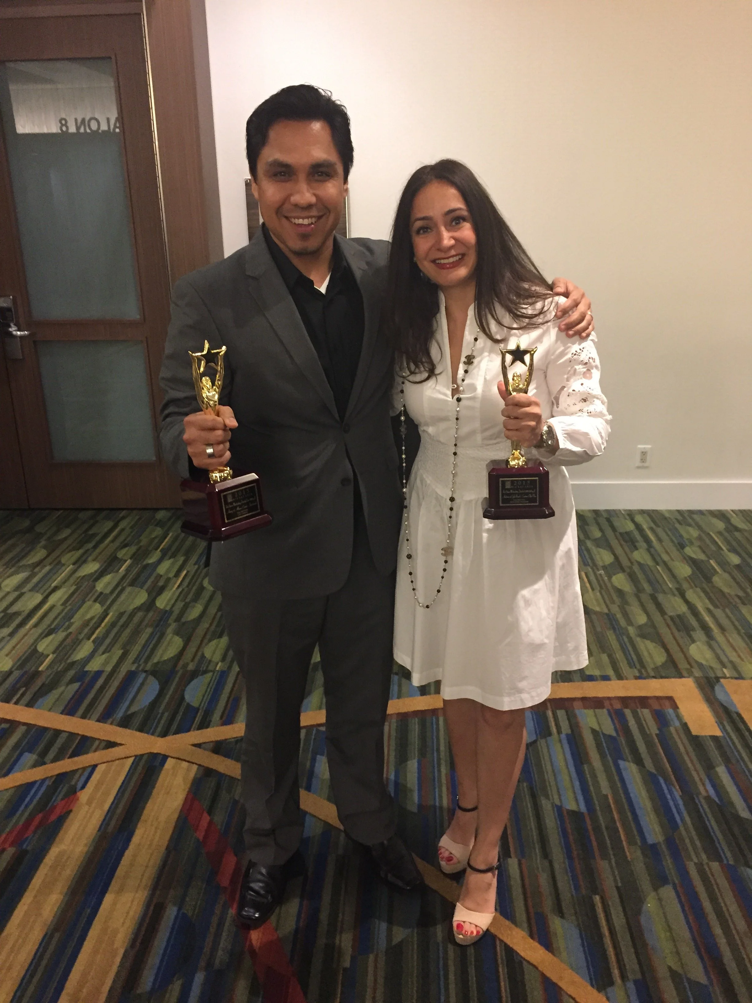 A man and woman smiling while holding awards, standing on a colorful patterned carpet, in an indoor setting with a plain wall and a wooden door in the background.