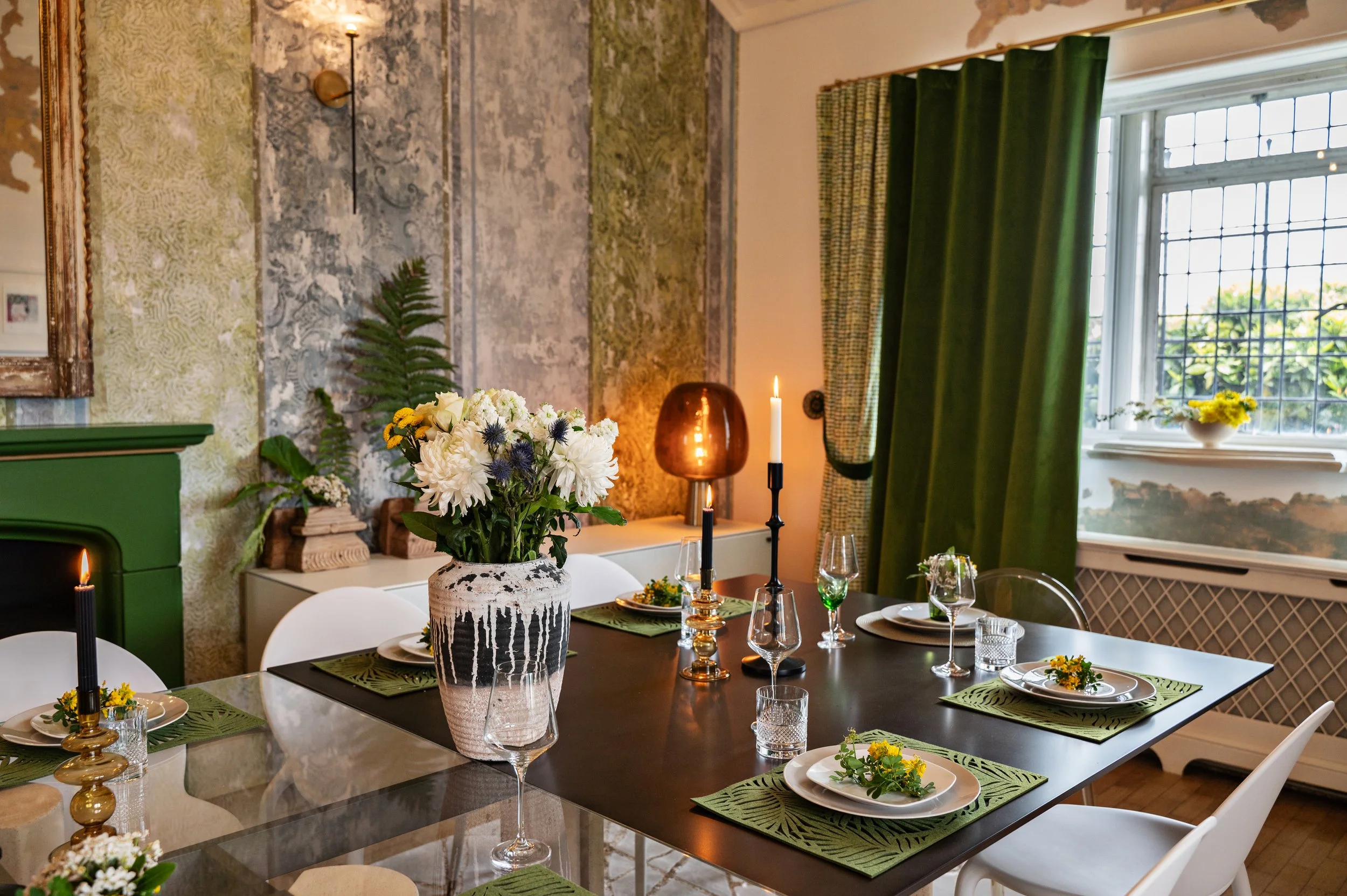 A dining room with a black table set with green placemats, white plates, glasses, and yellow flowers. A large bouquet of white flowers with blue accents is in a textured black and white vase in the center. The room has green curtains, a window, a patterned wall, and candles for decoration.