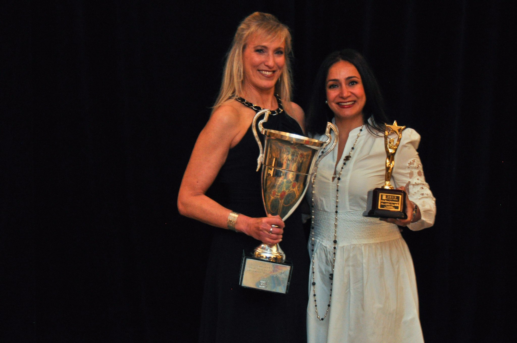 Two women smiling and holding trophies at an awards ceremony, standing against a black background.