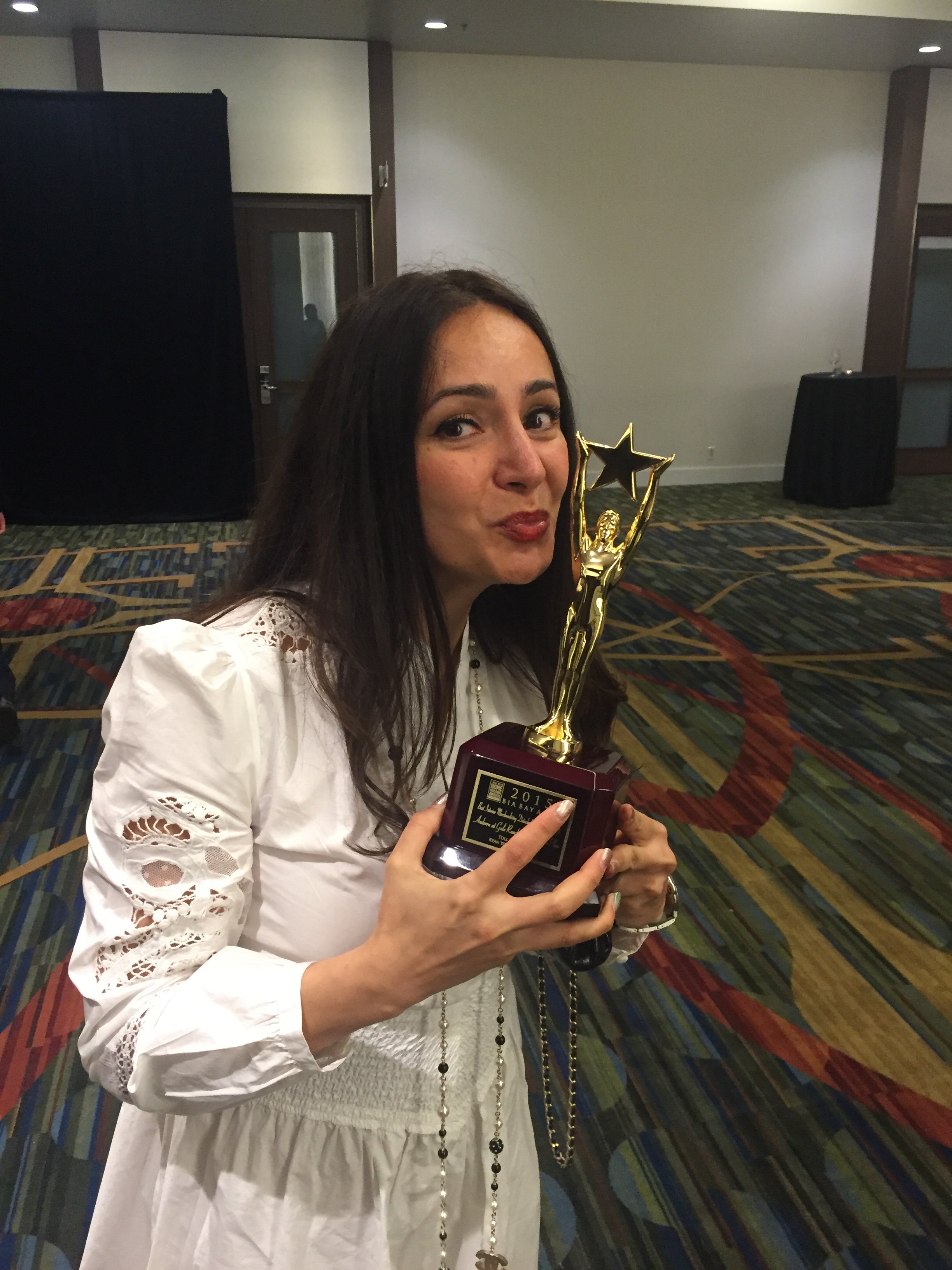 Woman with long dark hair holding a gold trophy with a star on top, inside a room with a decorative multicolored carpet and plain walls.