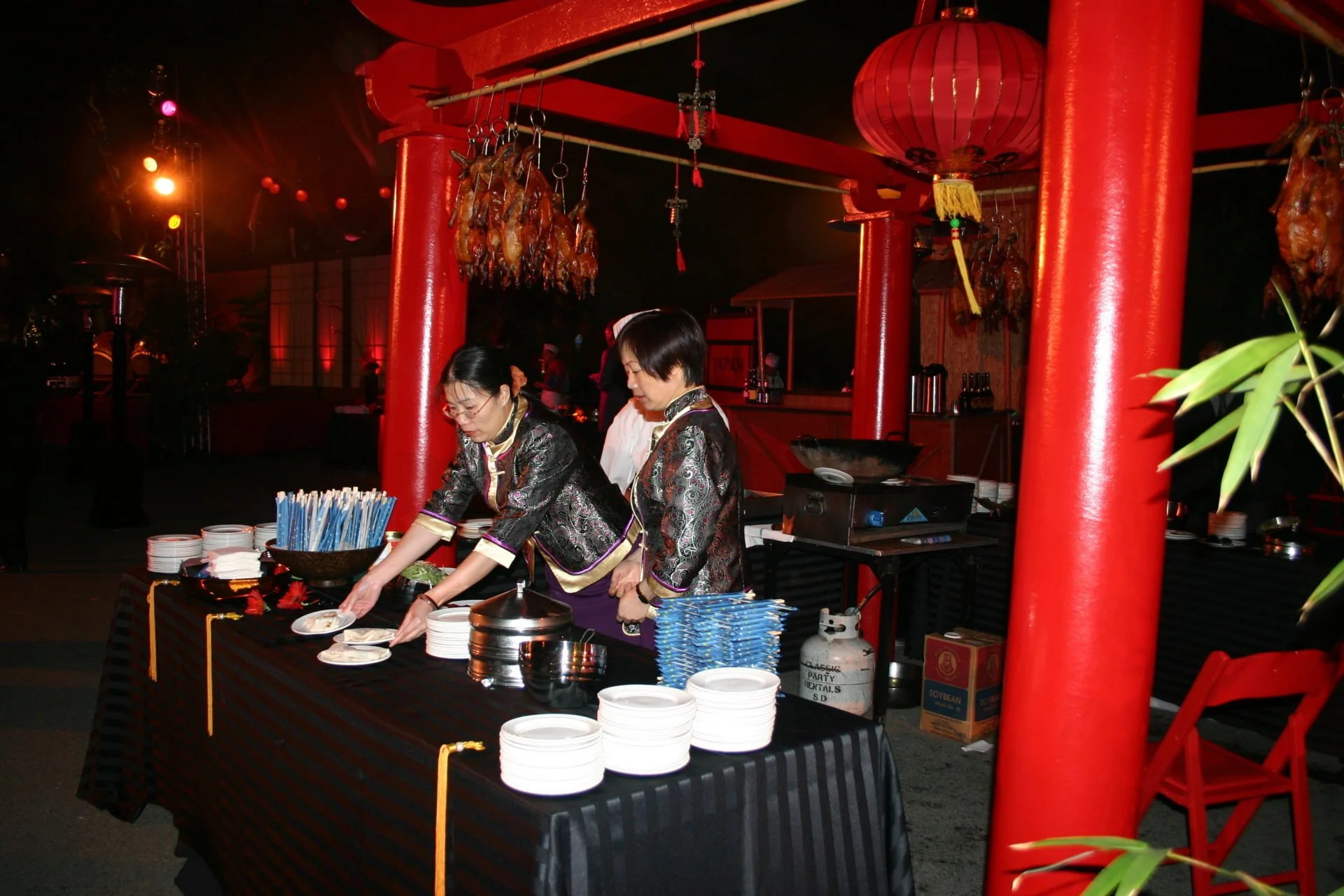 Two women in traditional Asian attire preparing food at a buffet table with plates, utensils, and food items in a setting decorated with red pillars and lanterns, likely during a cultural celebration or event.