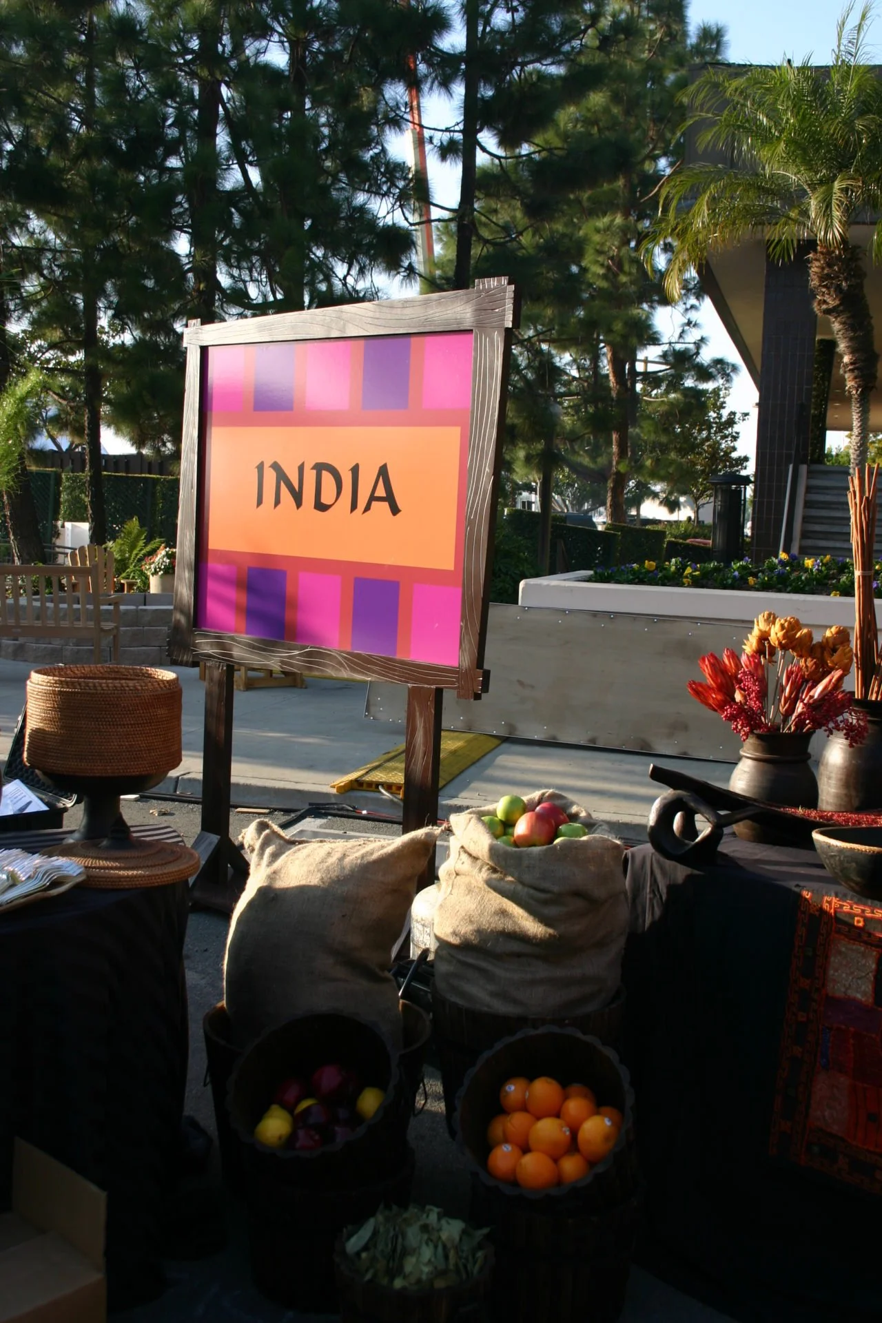 A colorful sign with the word 'INDIA' displayed in front of an outdoor market or festival scene with trees and plants, various baskets with fruits, and decorative items.