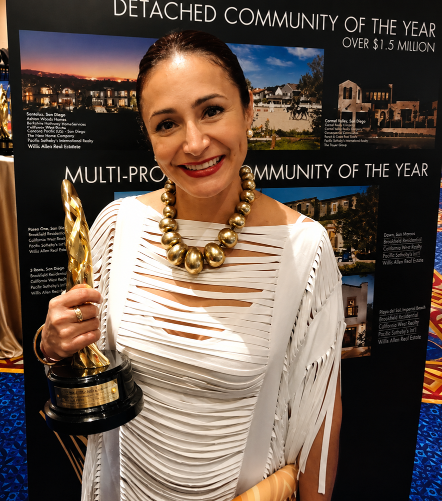 A woman smiling is holding a gold trophy in front of a black poster with images of upscale residential communities. She is wearing a white, draped dress with cutouts and a large gold bead necklace, and has pearl earrings.