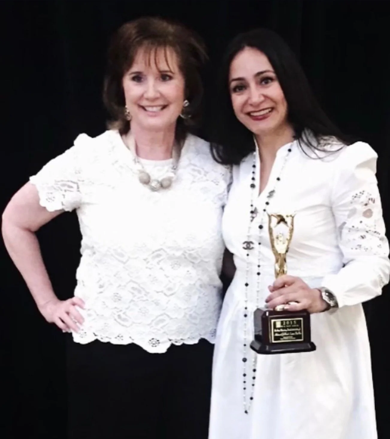 Two women in white clothing standing together. One woman holds an award.