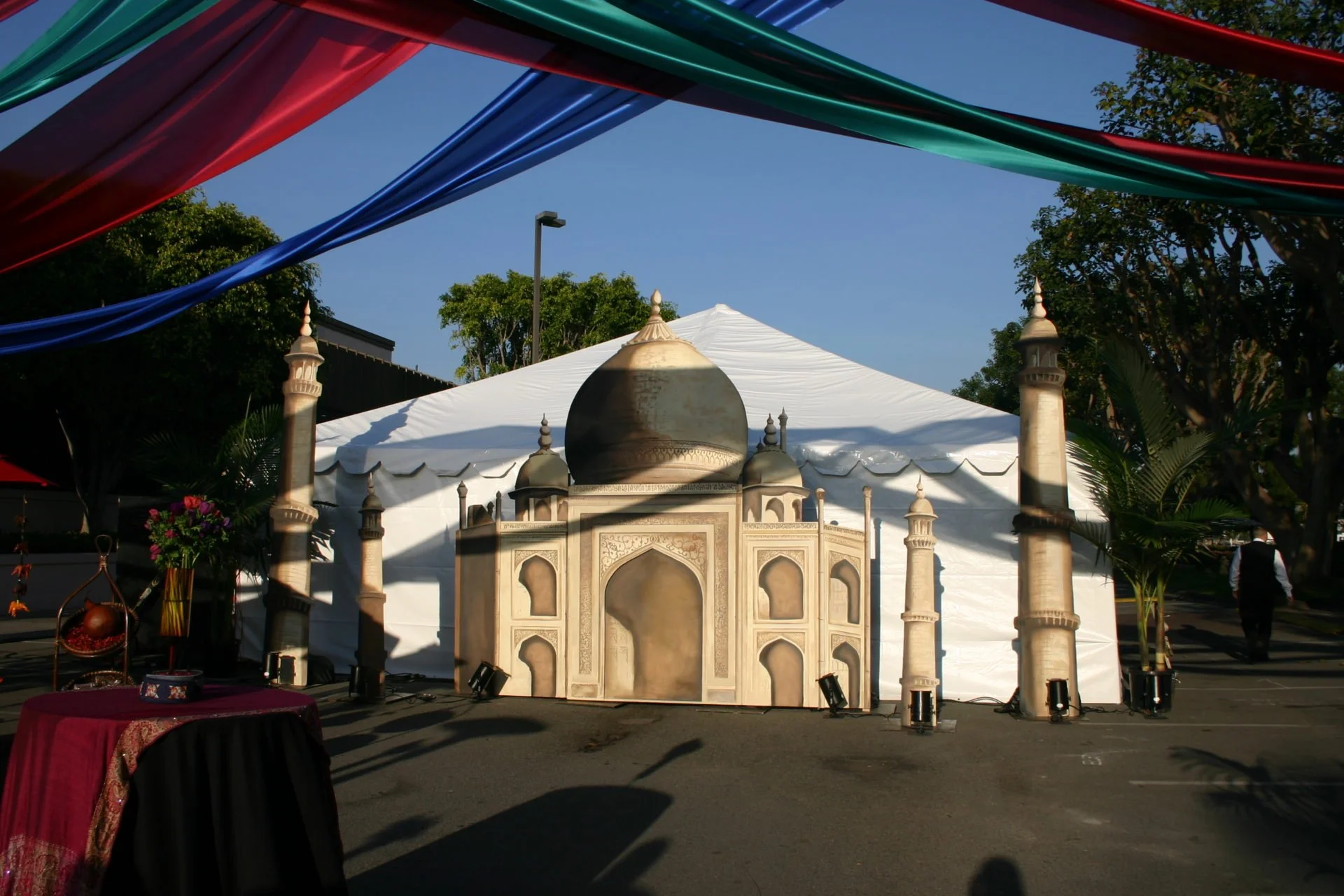A decorated outdoor event space with a large white tent, a structure resembling the Taj Mahal, and colorful fabric drapes overhead. There are trees and a clear blue sky in the background.