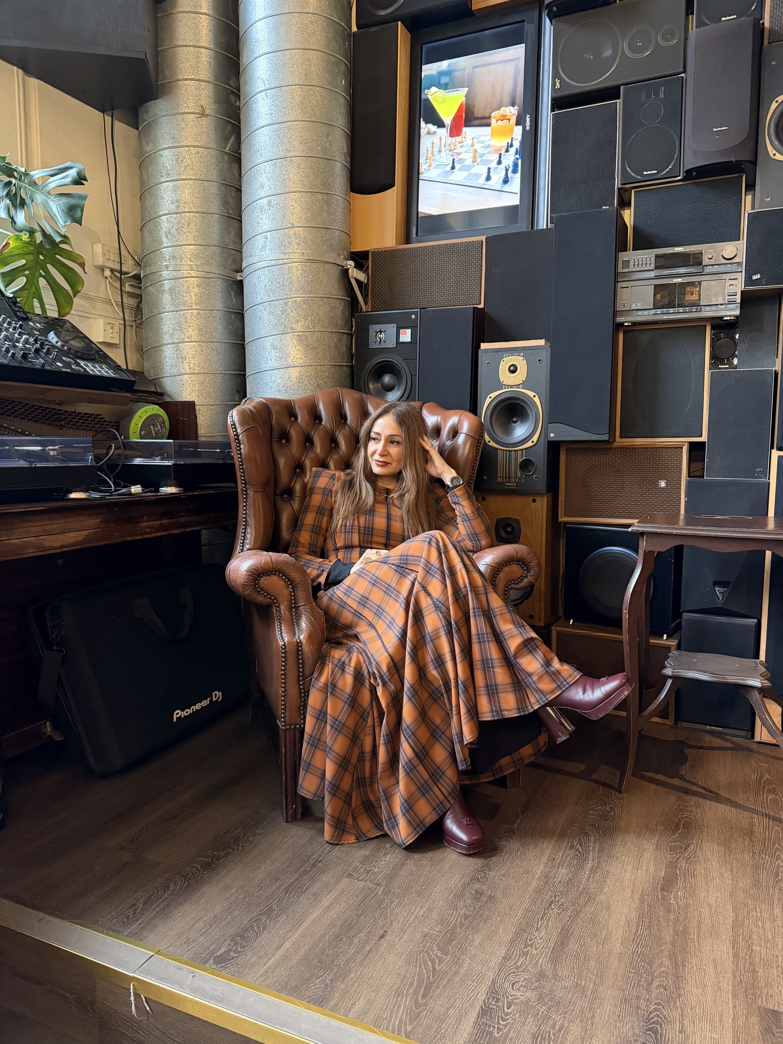 A woman sitting on a brown leather armchair in a room filled with audio equipment and speakers, with a woman wearing a plaid suit, purple shoes, and a watch.