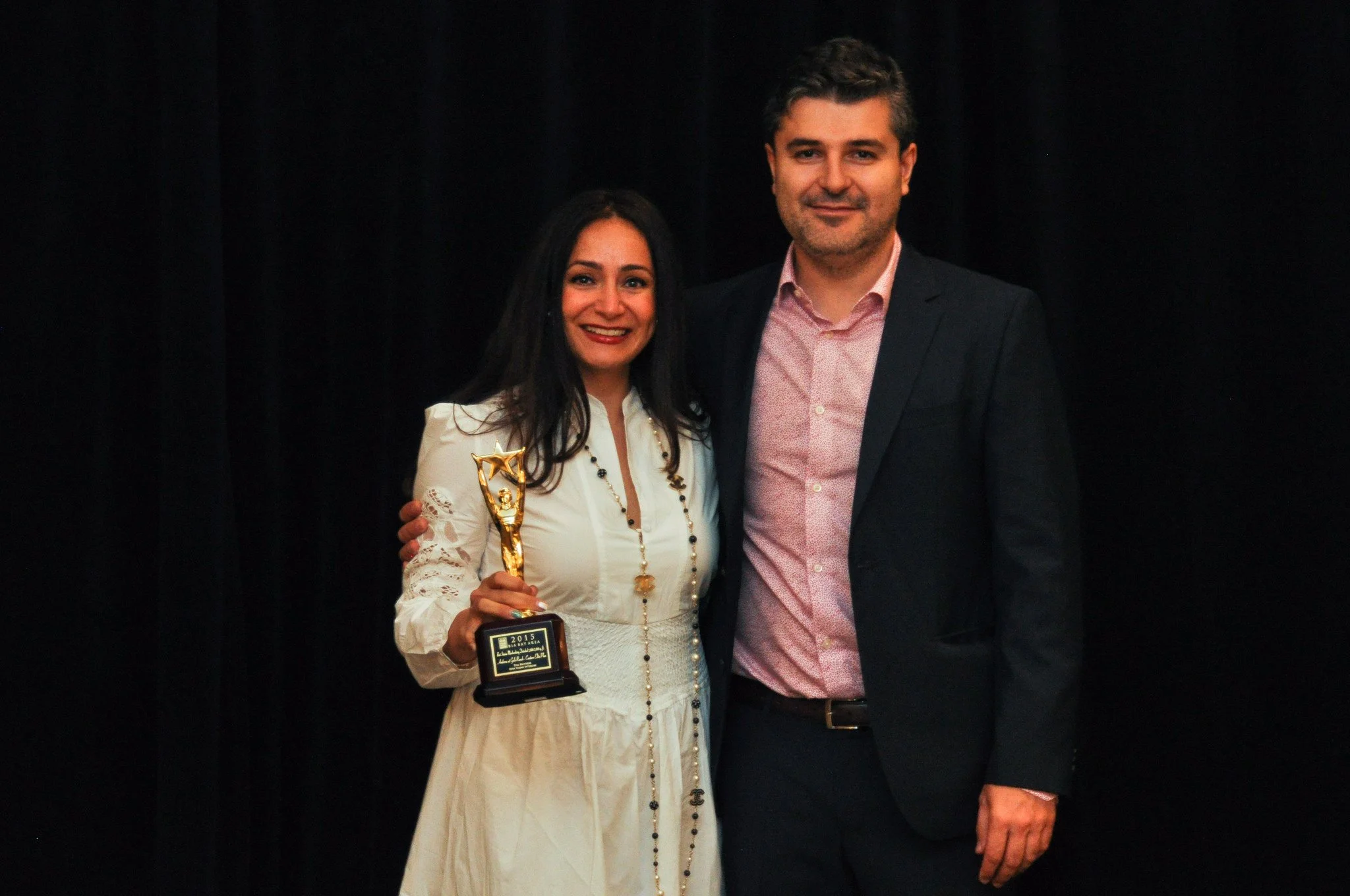 A woman holding a trophy and smiling, standing next to a man in a suit. The woman is wearing a white dress with lace sleeves and a necklace, and has long dark hair. The man is wearing a pink shirt and a dark blazer. They are posed against a dark back