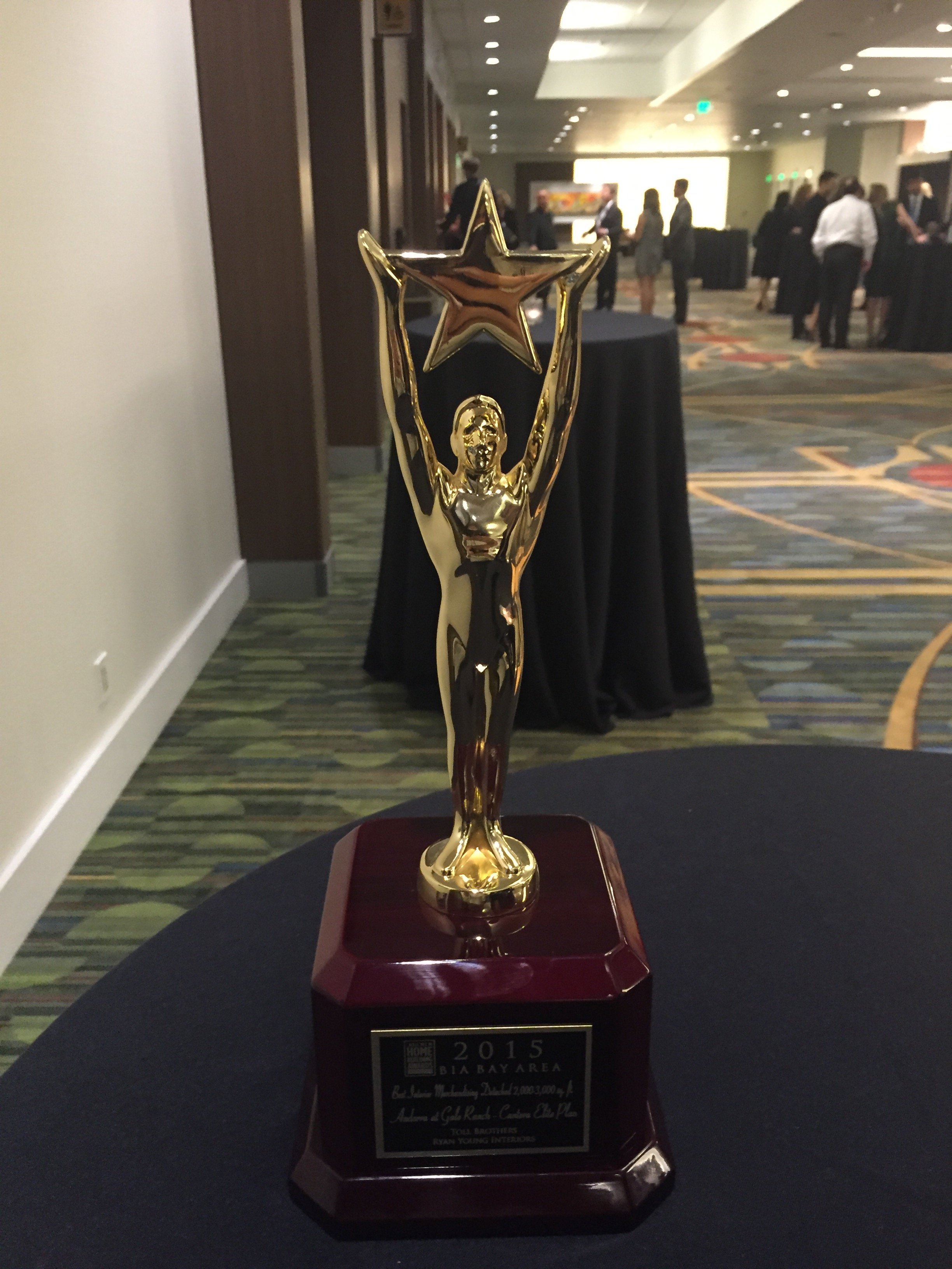 Gold trophy with a star and a figure holding it up, placed on a dark table in a large, well-lit conference room with people in the background.