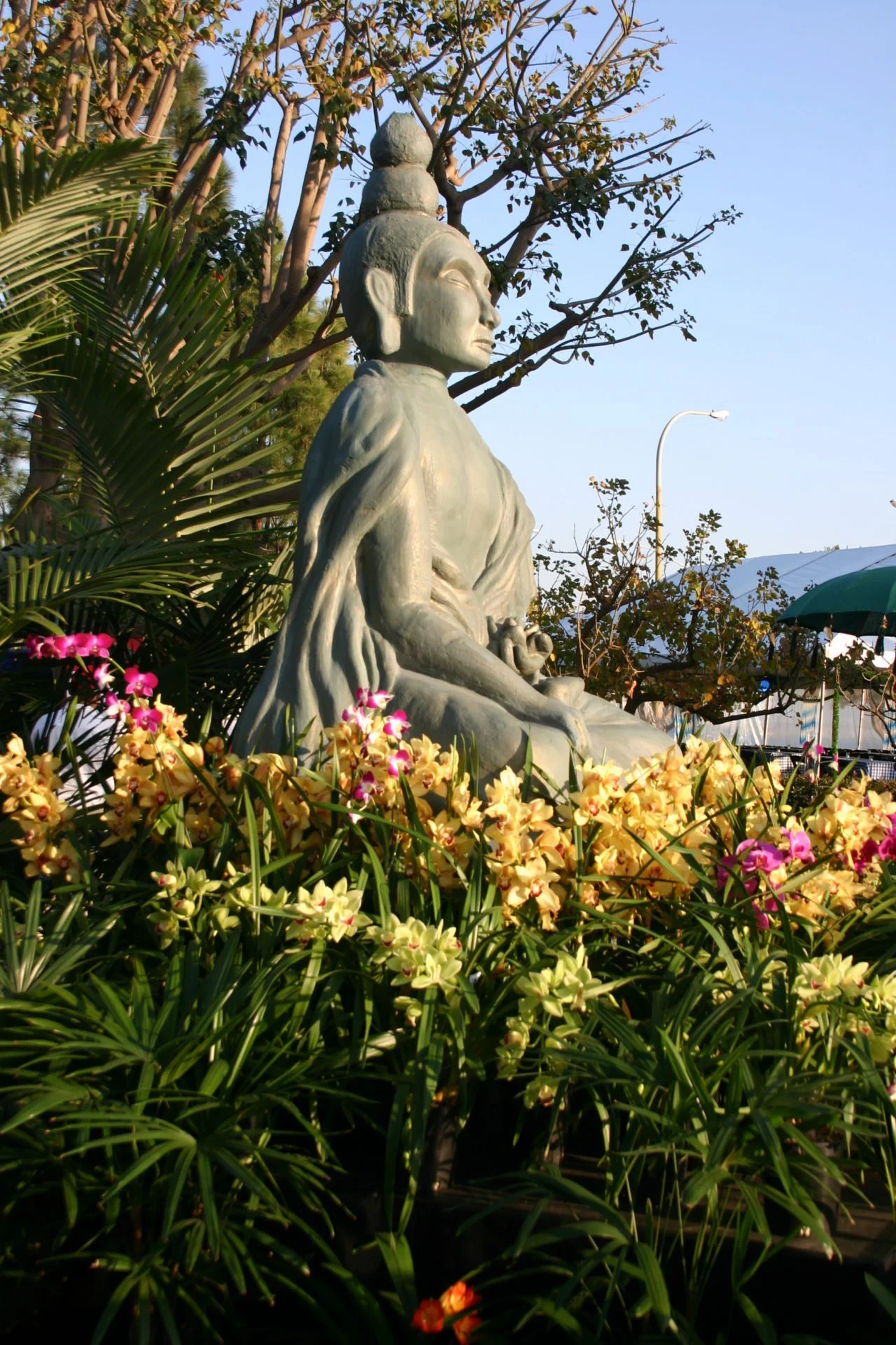 Statue of a seated figure with a headdress, surrounded by colorful flowers and lush greenery, under a clear blue sky.