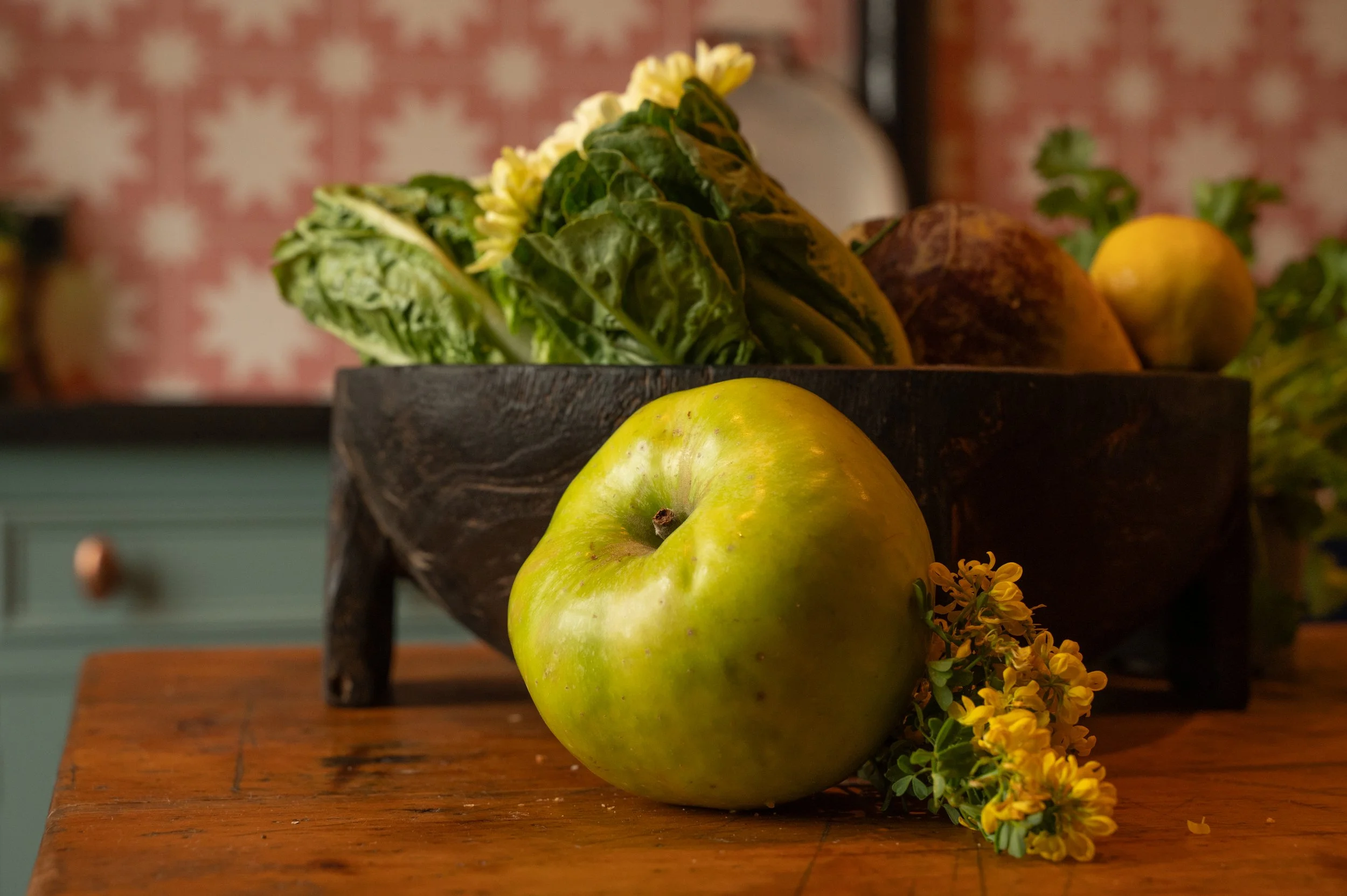 A green apple, a wooden bowl of leafy greens and other vegetables, and yellow flowers on a wooden table in a rustic interior.