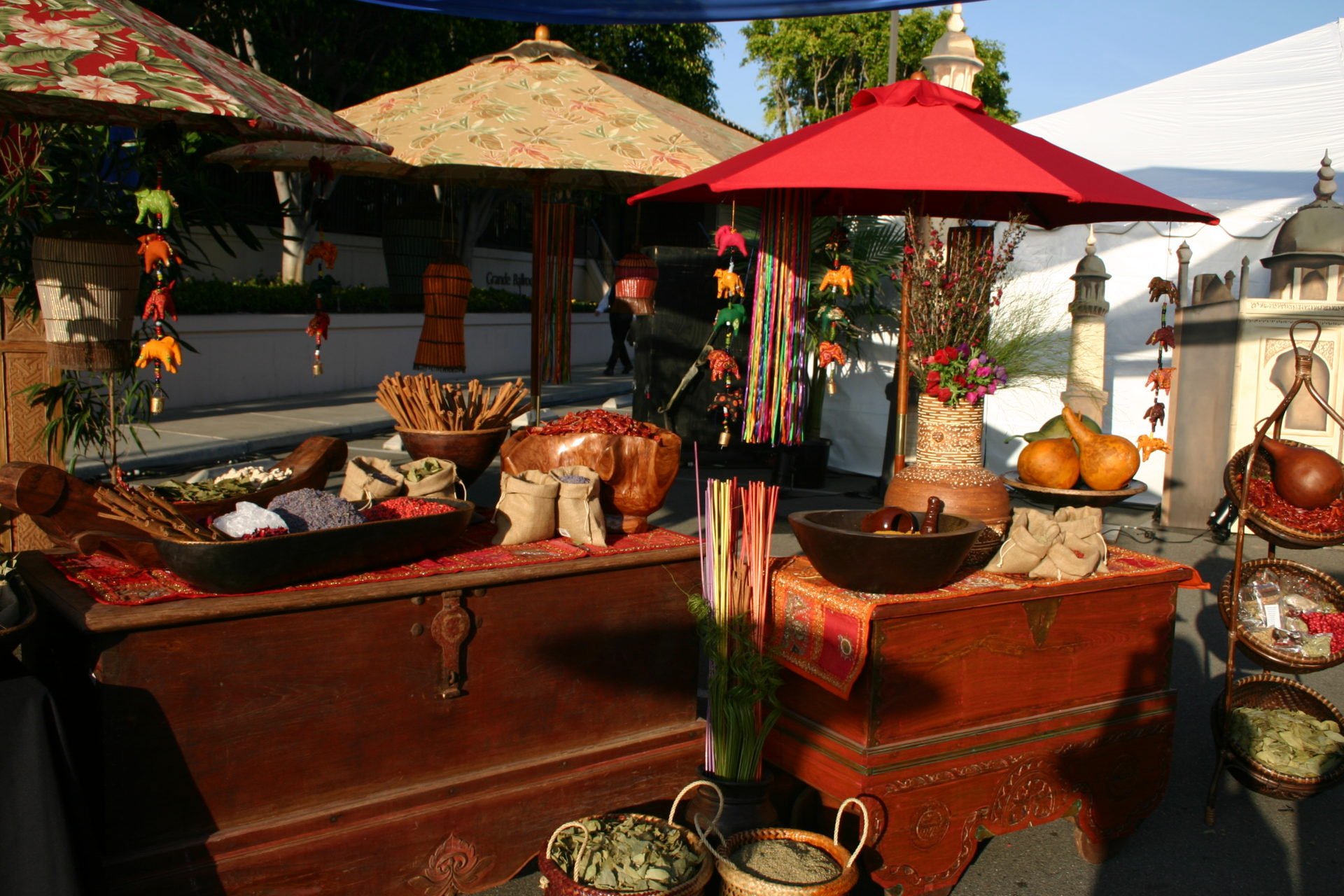 Outdoor market stall with various colorful textiles, bags, and dried herbs under a bright red umbrella, with decorative hanging ornaments, plants, and pottery around.