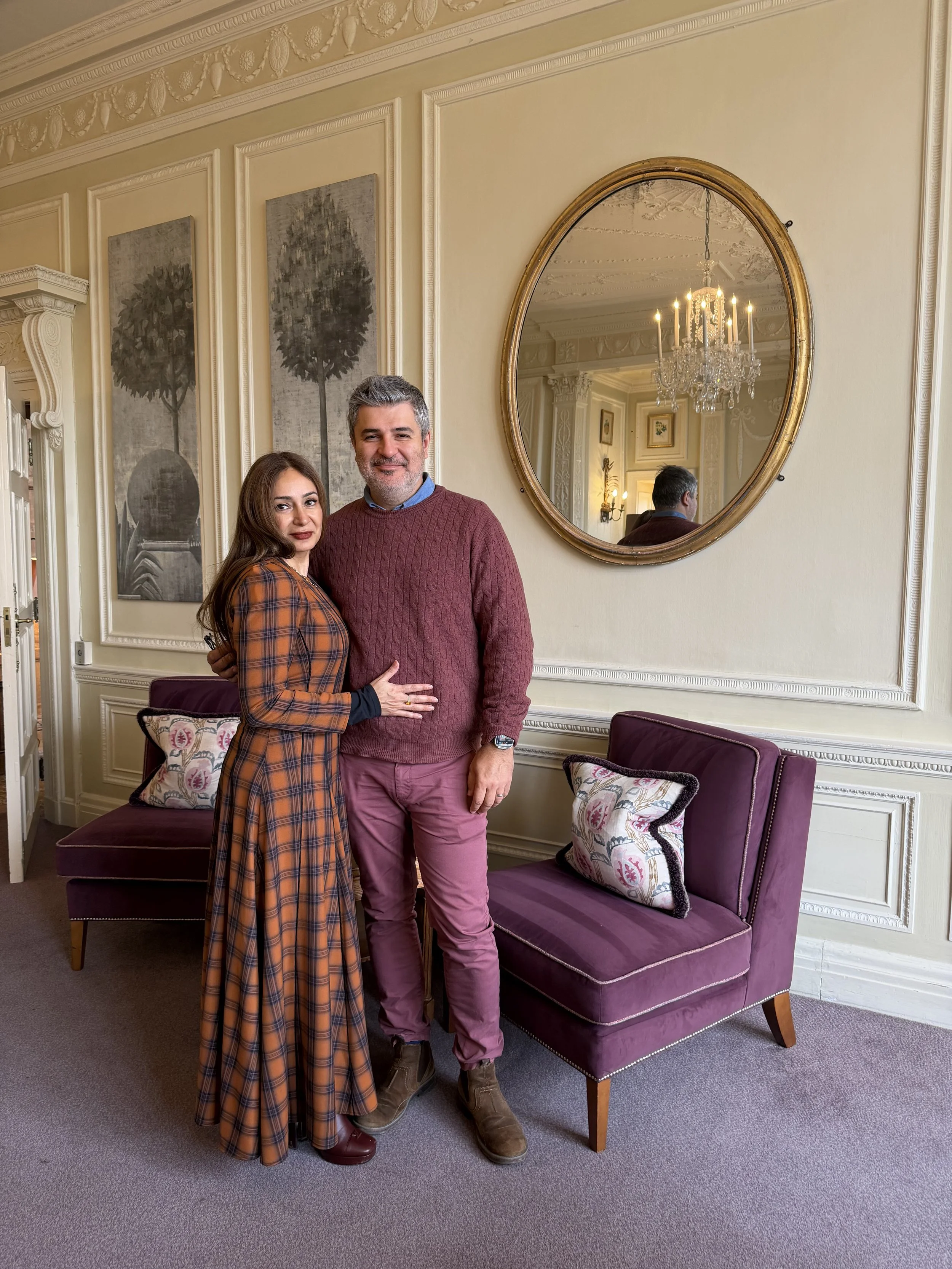 A woman and a man standing close together on a gray carpeted floor in a living room, with their arms around each other, smiling at the camera. The woman is wearing a long plaid dress and the man is wearing a maroon sweater, pink pants, and brown boot