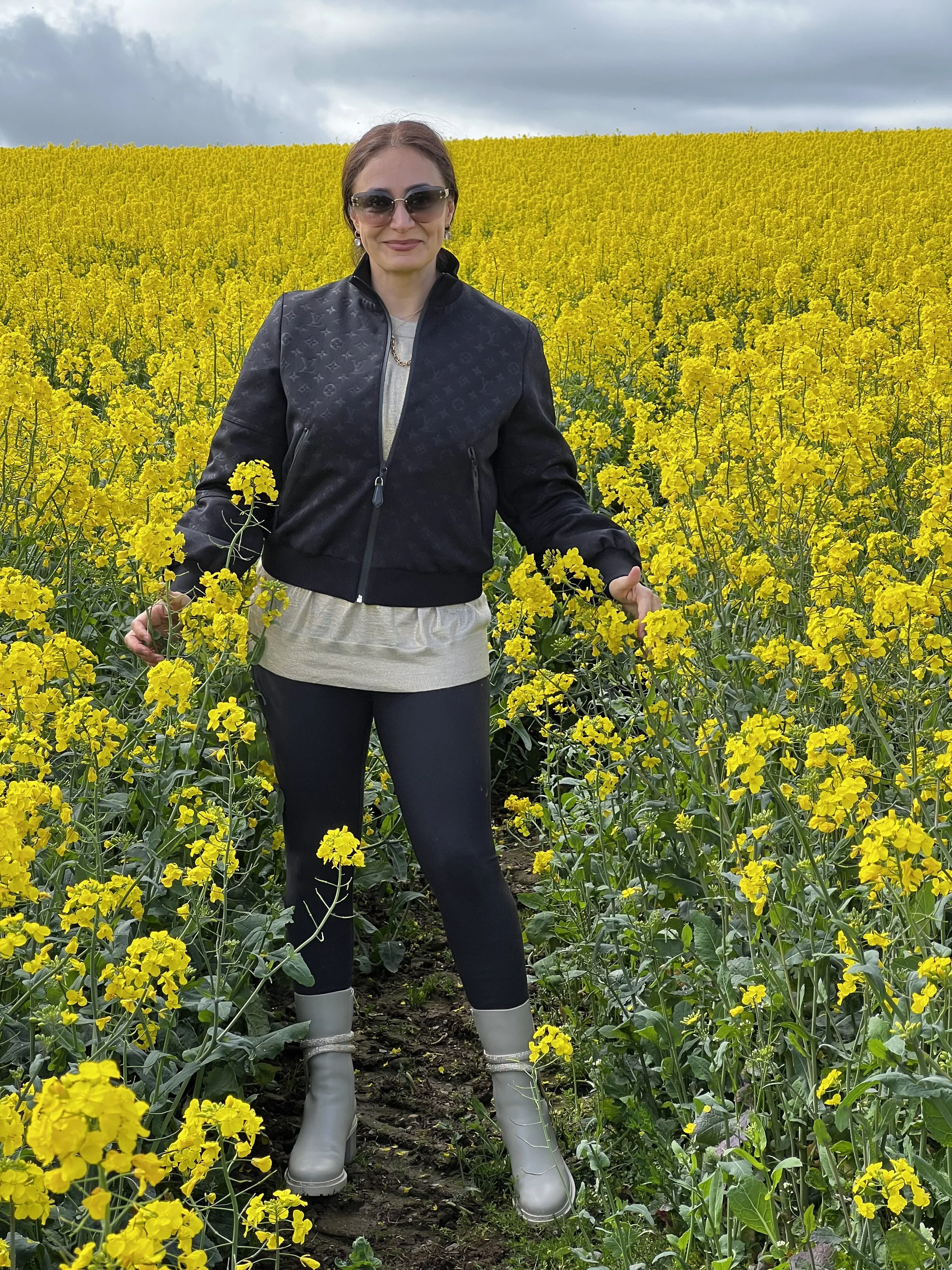 A woman standing in a field of bright yellow flowers, wearing sunglasses, a black jacket, beige top, black pants, and grey rubber boots, with a cloudy sky above.
