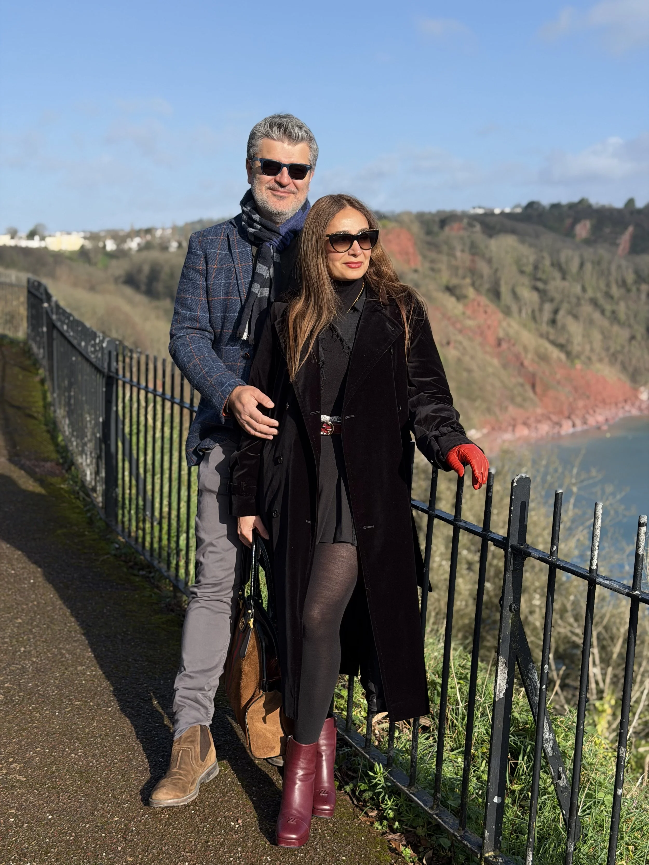 Two people, a man and a woman, stand next to a metal fence on a walkway overlooking a scenic landscape of cliffs, ocean, and a partly cloudy sky, during daytime. They are dressed warmly and wearing sunglasses.