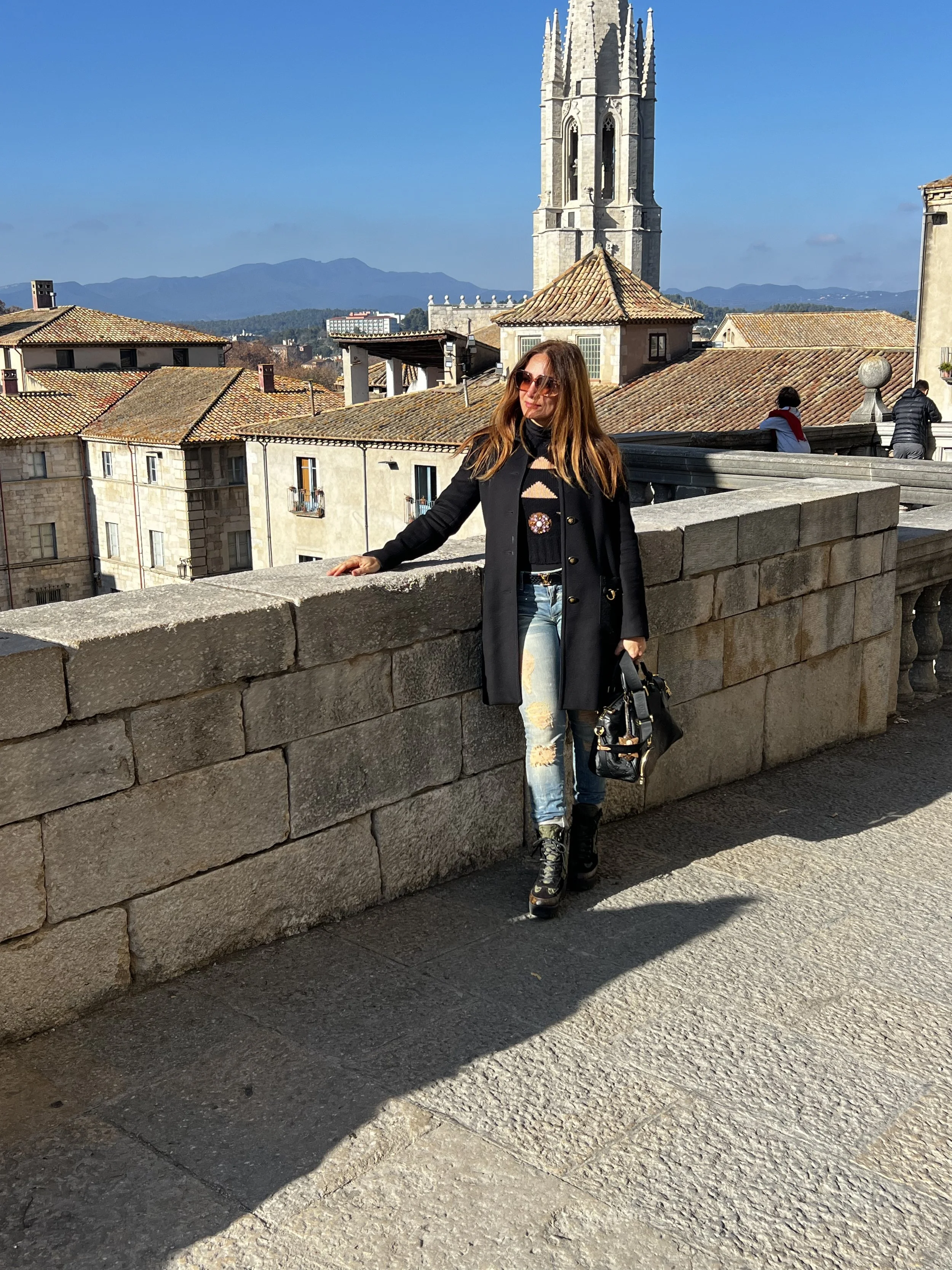 A woman wearing sunglasses, a black coat, ripped jeans, and boots, is leaning on a stone wall in front of a historic cityscape with a tall church tower and tiled rooftops, under a clear blue sky with mountains in the background.