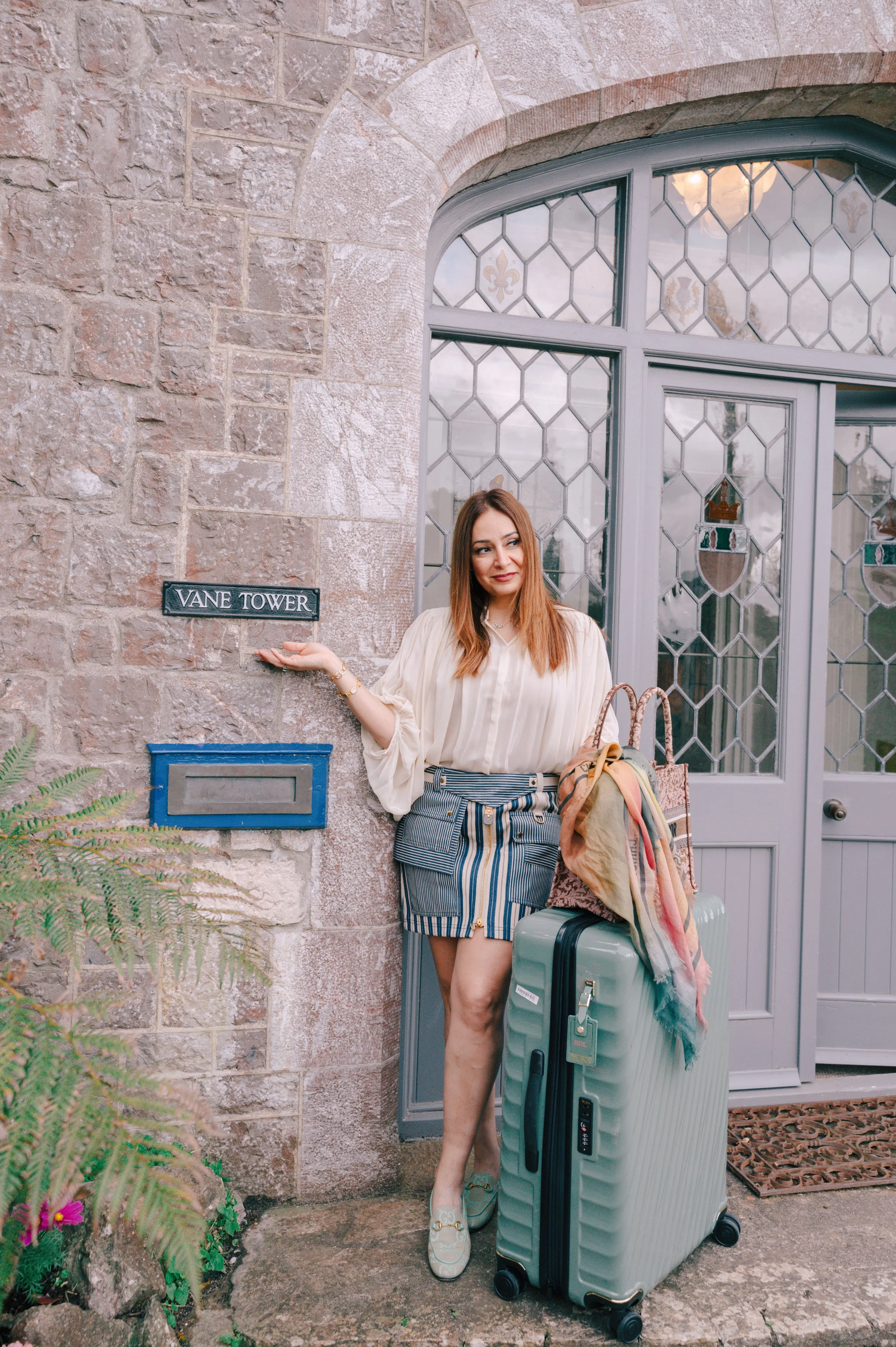 A woman with long brown hair standing outside a stone building with a window and a door, holding a sign that says 'Vane Tower'. She is wearing a cream-colored blouse, a striped skirt, and sneakers. She has a large suitcase, a handbag, and a scarf wit