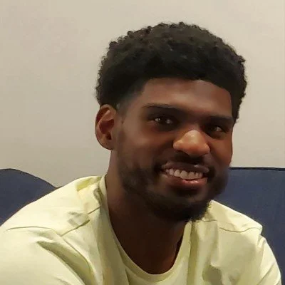 A man with dark curly hair and a beard smiling at the camera, wearing a light-colored shirt, sitting on a blue couch against a plain light-colored wall.