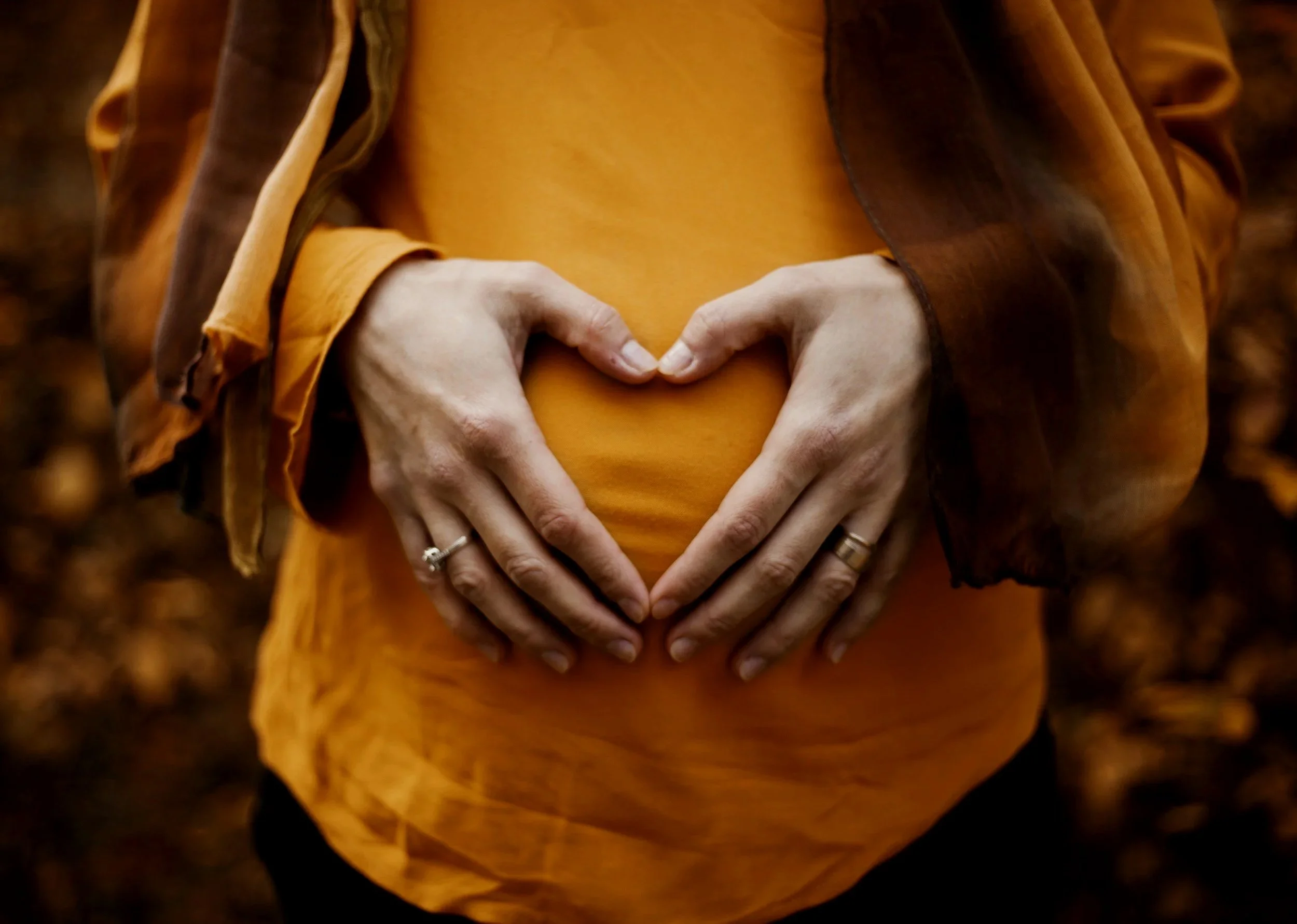 Person wearing an orange shirt and brown jacket forming a heart shape with hands over chest.