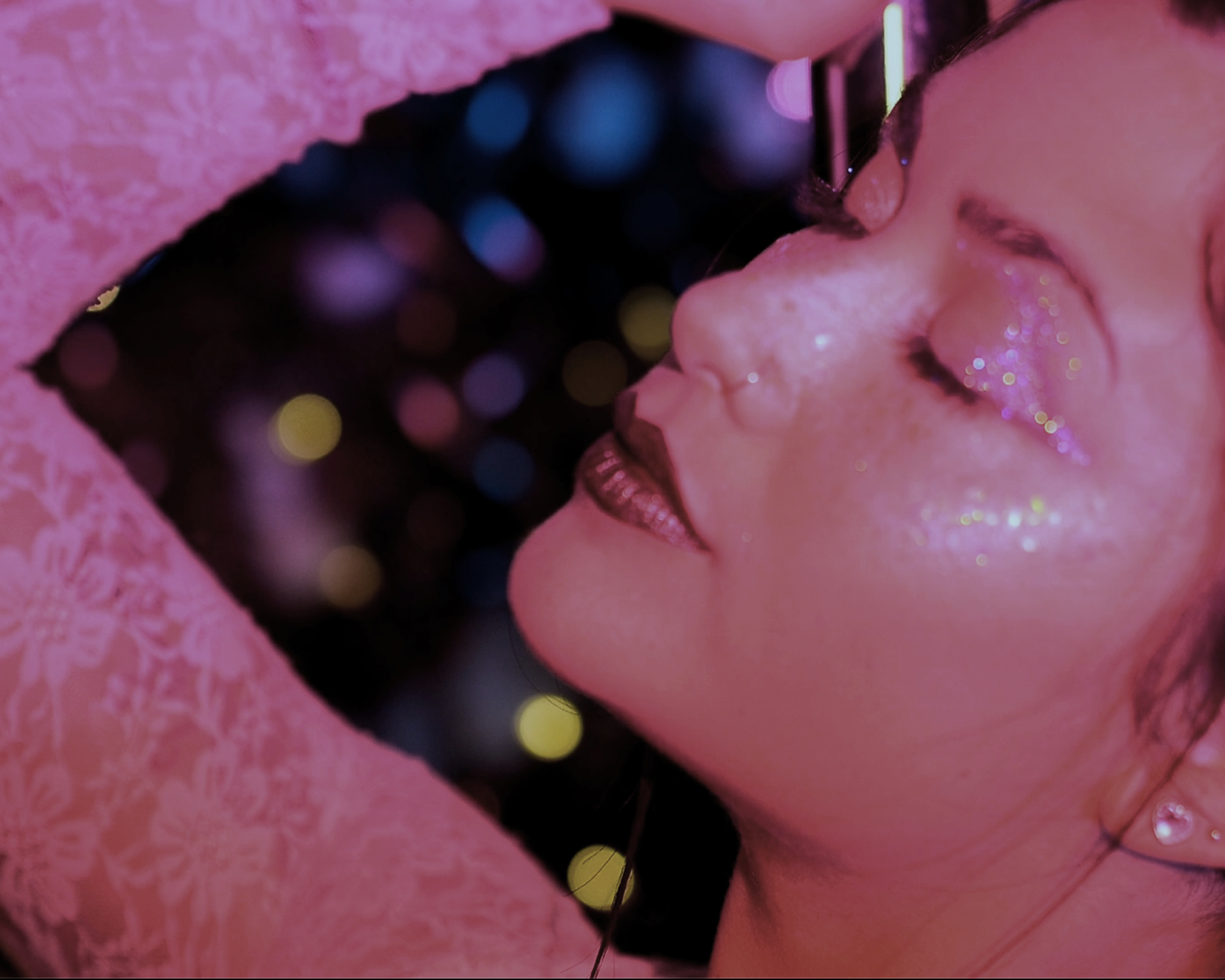Close-up of a woman with glittery makeup and earrings, covered by a pink lace fabric, with colorful bokeh lights in the background.