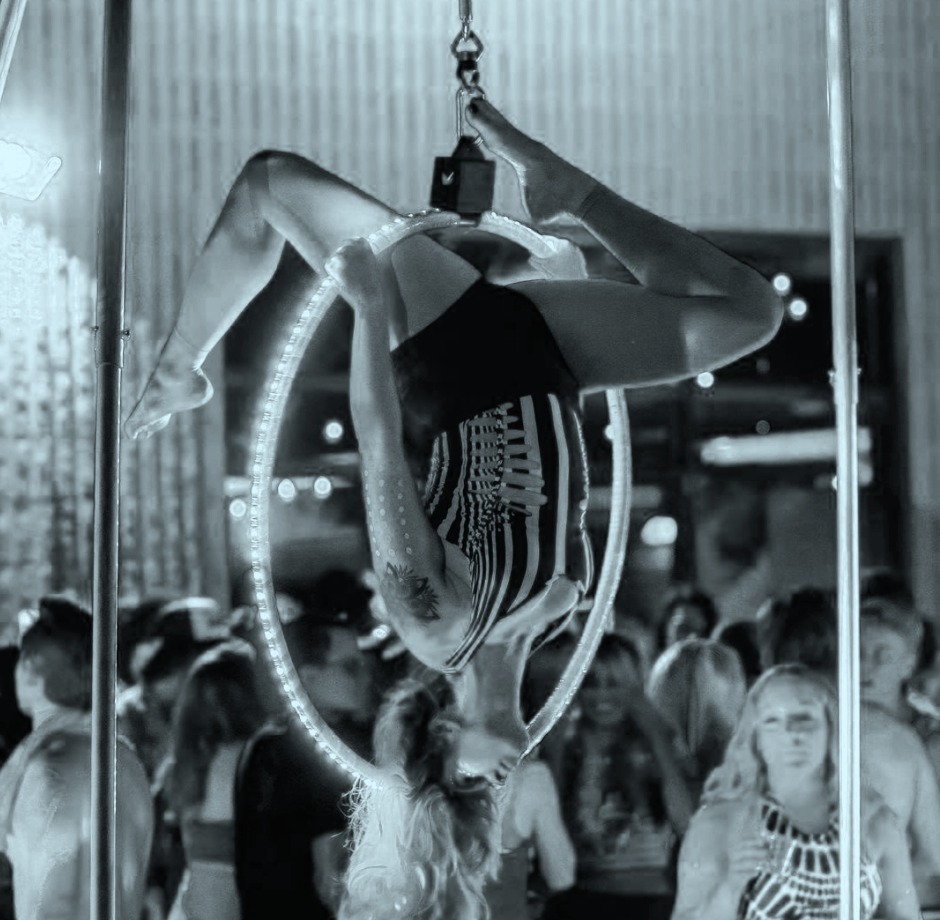 A female aerialist performing an upside-down act on a hoop at a crowded event, with spectators watching in the background.
