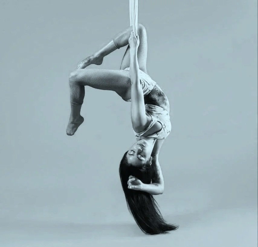 A woman performing an aerial acrobatic pose hanging upside down with long hair hanging towards the ground.