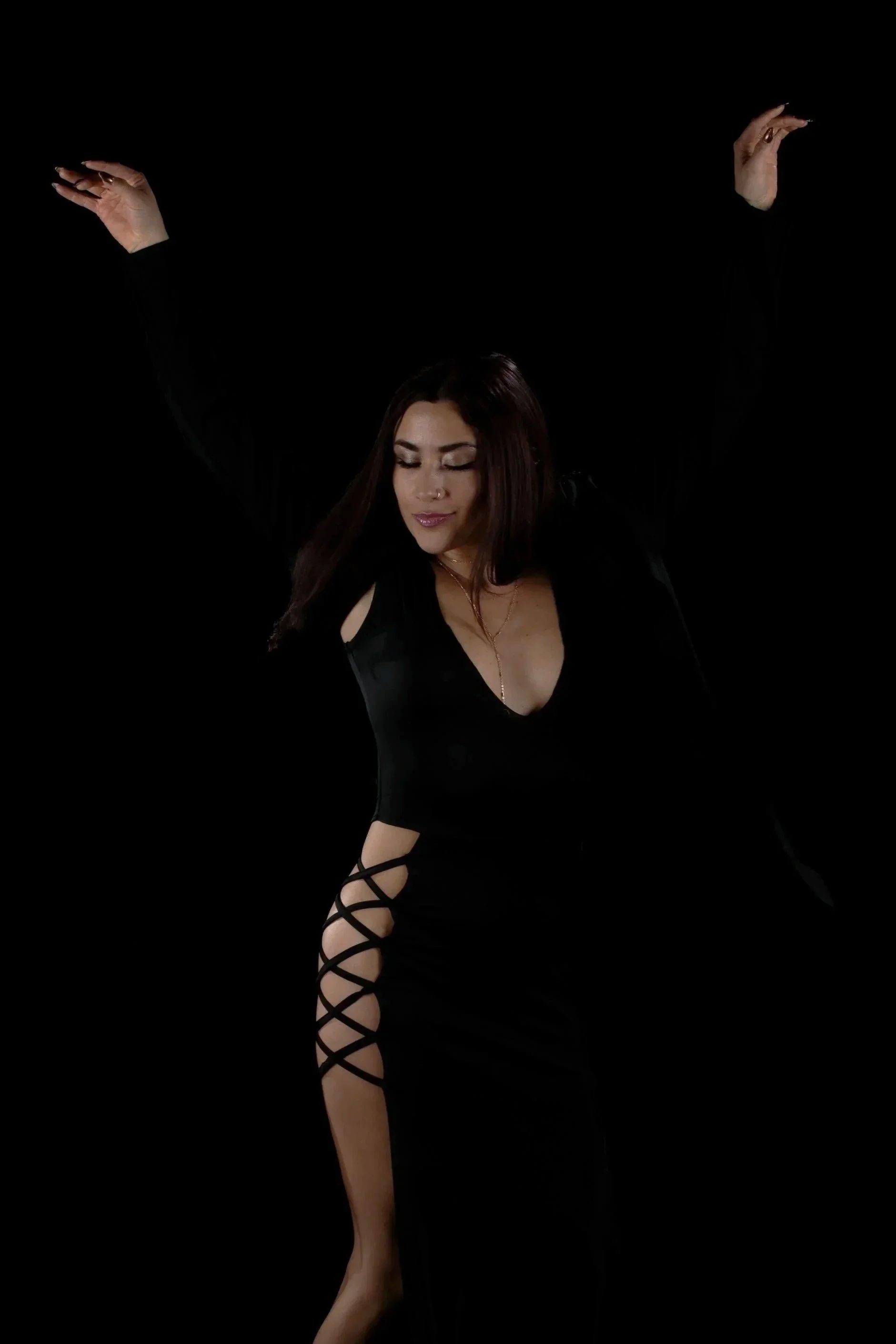 A woman with long brown hair wearing a black dress with cutout shoulder details and lace-up stockings, posing against a black background.
