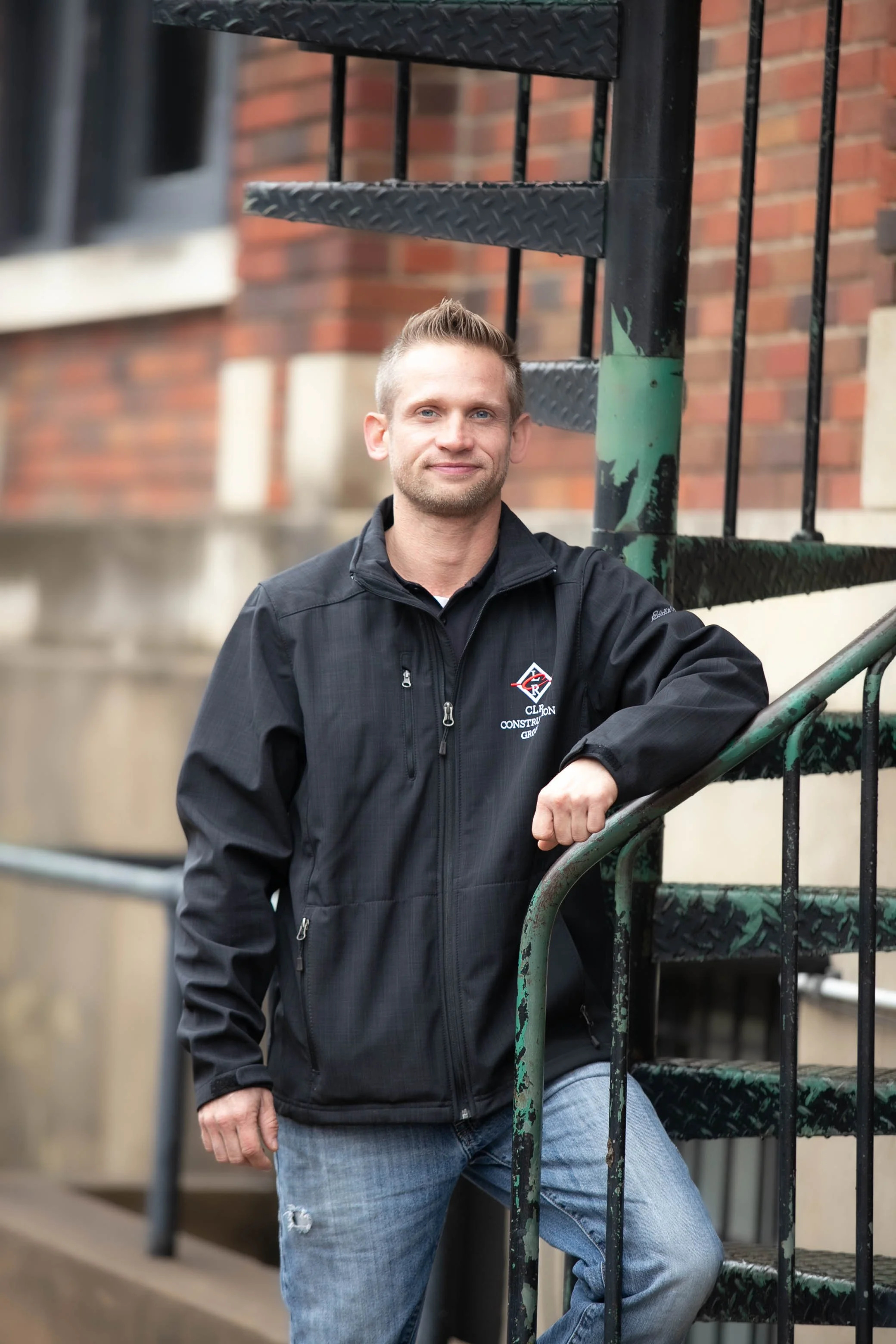 Man wearing a black jacket with a logo, standing outdoors by a metal staircase, with a brick building in the background.