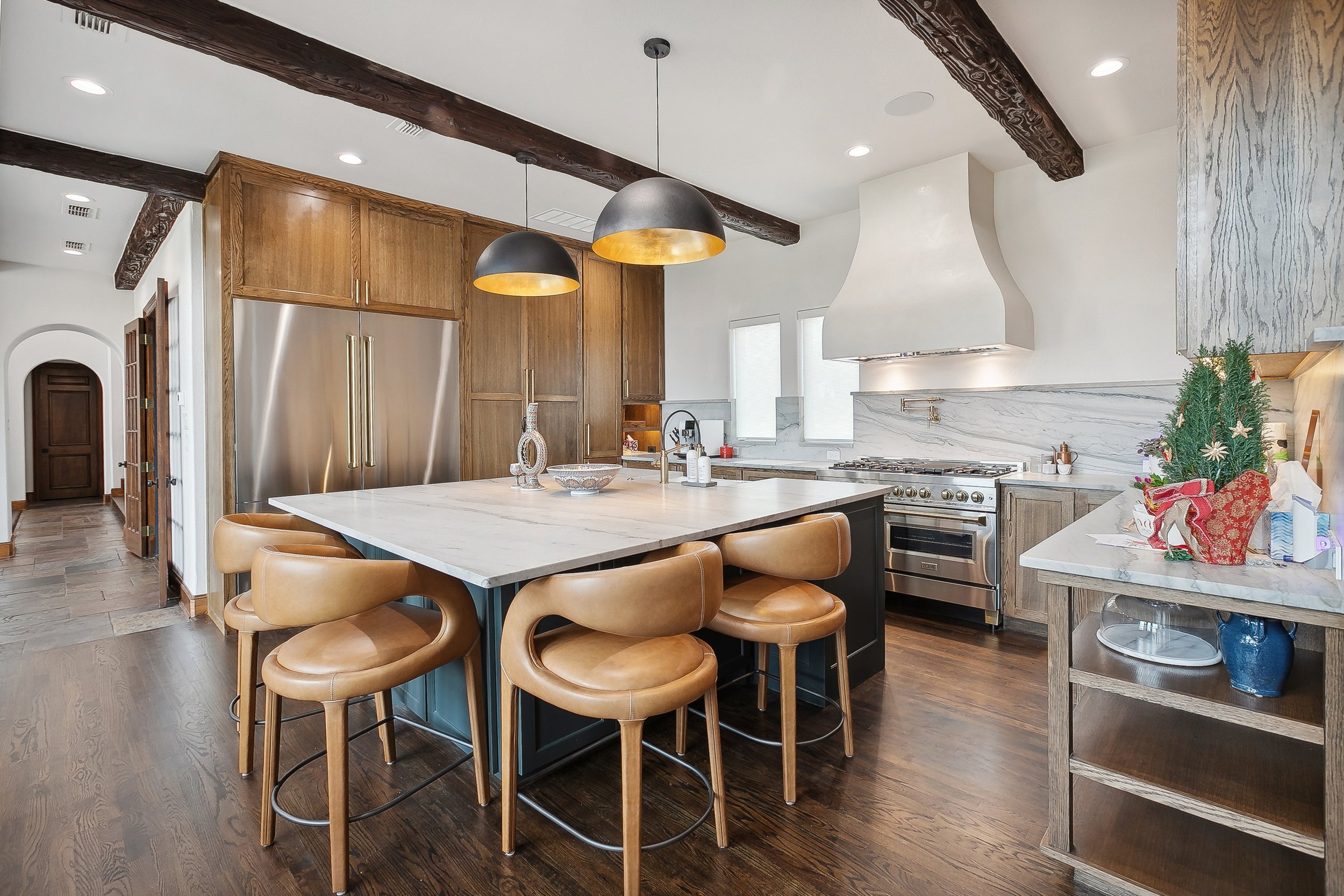 A modern kitchen with wooden cabinets, stainless steel refrigerator, marble countertops, a white hood above the stove, and ceiling beams. There is a marble island with tan leather chairs and pendant lights hanging above.