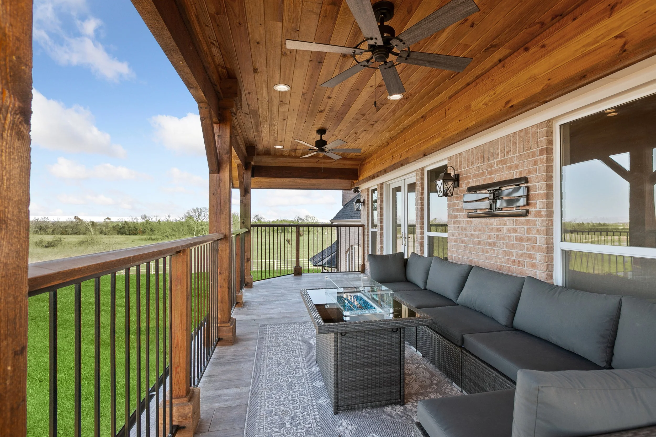 Covered balcony with wooden ceiling, ceiling fans, brick wall, large windows, outdoor seating, and a view of green fields and blue sky.