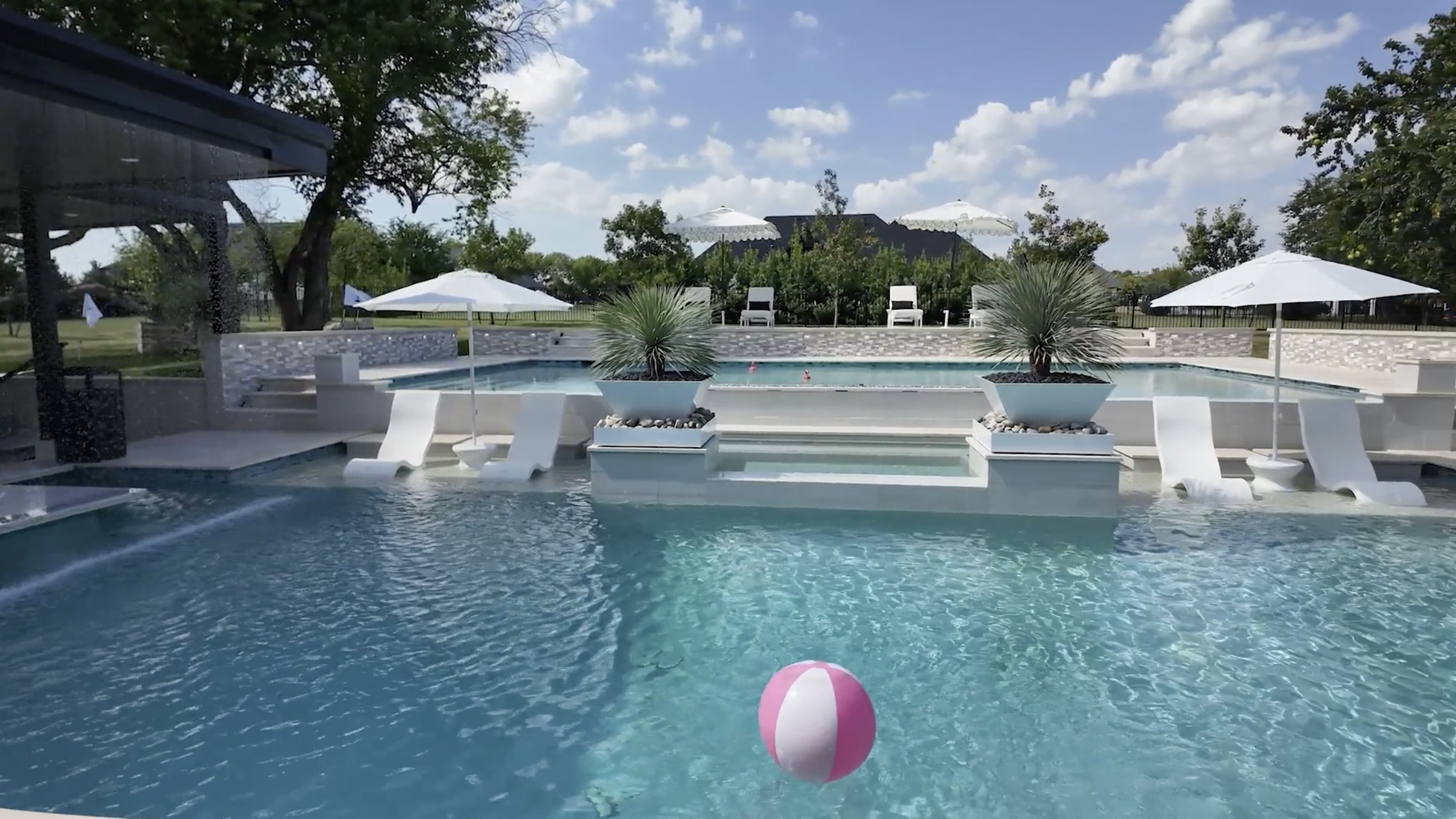 A backyard swimming pool with white lounge chairs and umbrellas, surrounded by greenery and trees under a partly cloudy sky.