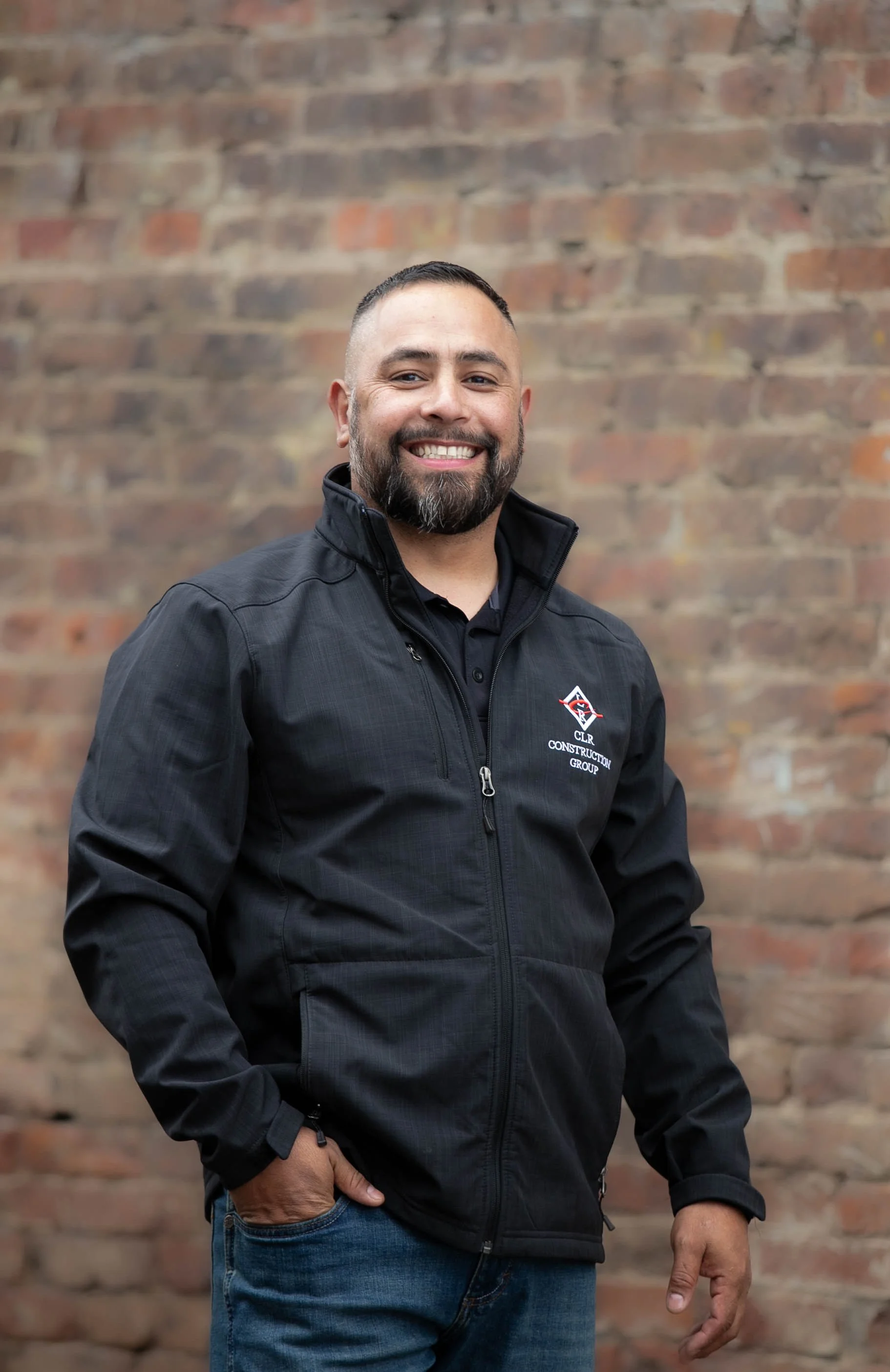 A smiling man with short dark hair and a beard, wearing a black jacket with a logo, standing in front of a brick wall.