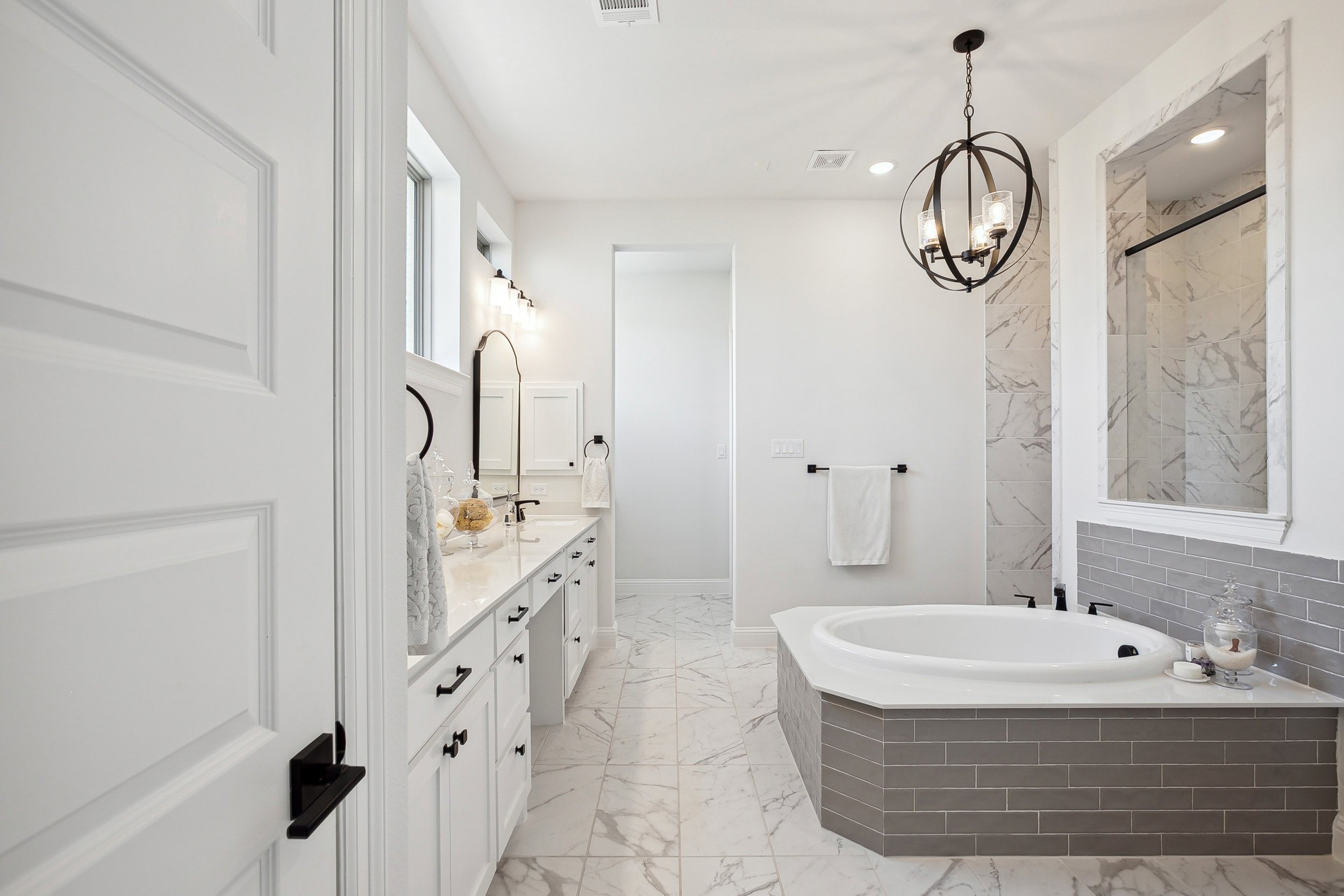 A modern, bright bathroom with white walls and marble flooring. It features a white vanity with black handles, a bathtub with a gray tiled exterior, a chandelier, and a walk-in shower with marble tiles.