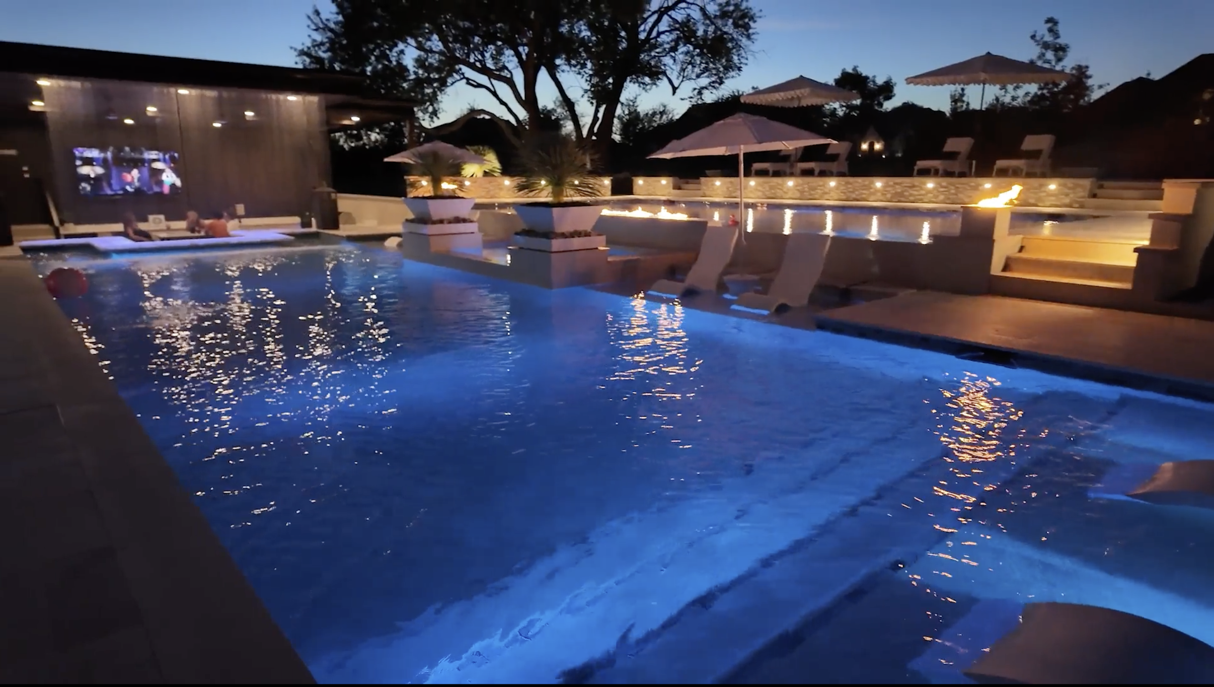 A nighttime view of a swimming pool with blue lighting, surrounded by lounge chairs and umbrellas, with a fire feature and a large tree in the background.