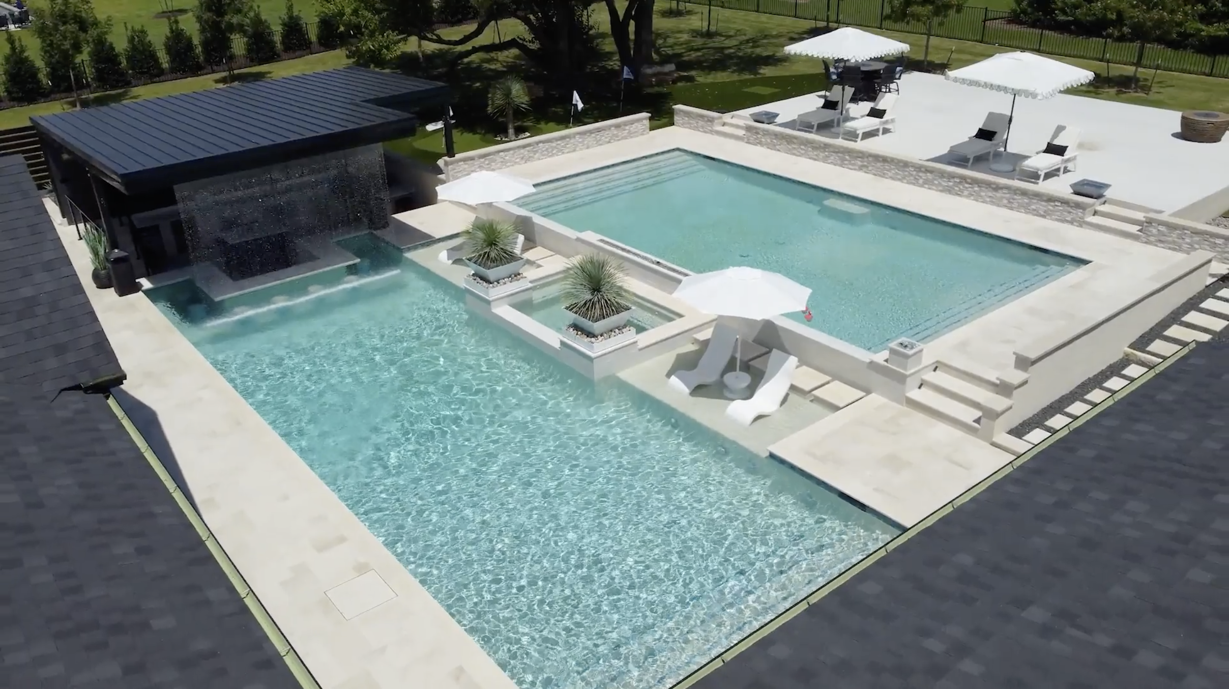 A luxurious backyard with a modern swimming pool and spa, surrounded by white patio furniture, umbrellas, and a manicured lawn with trees.