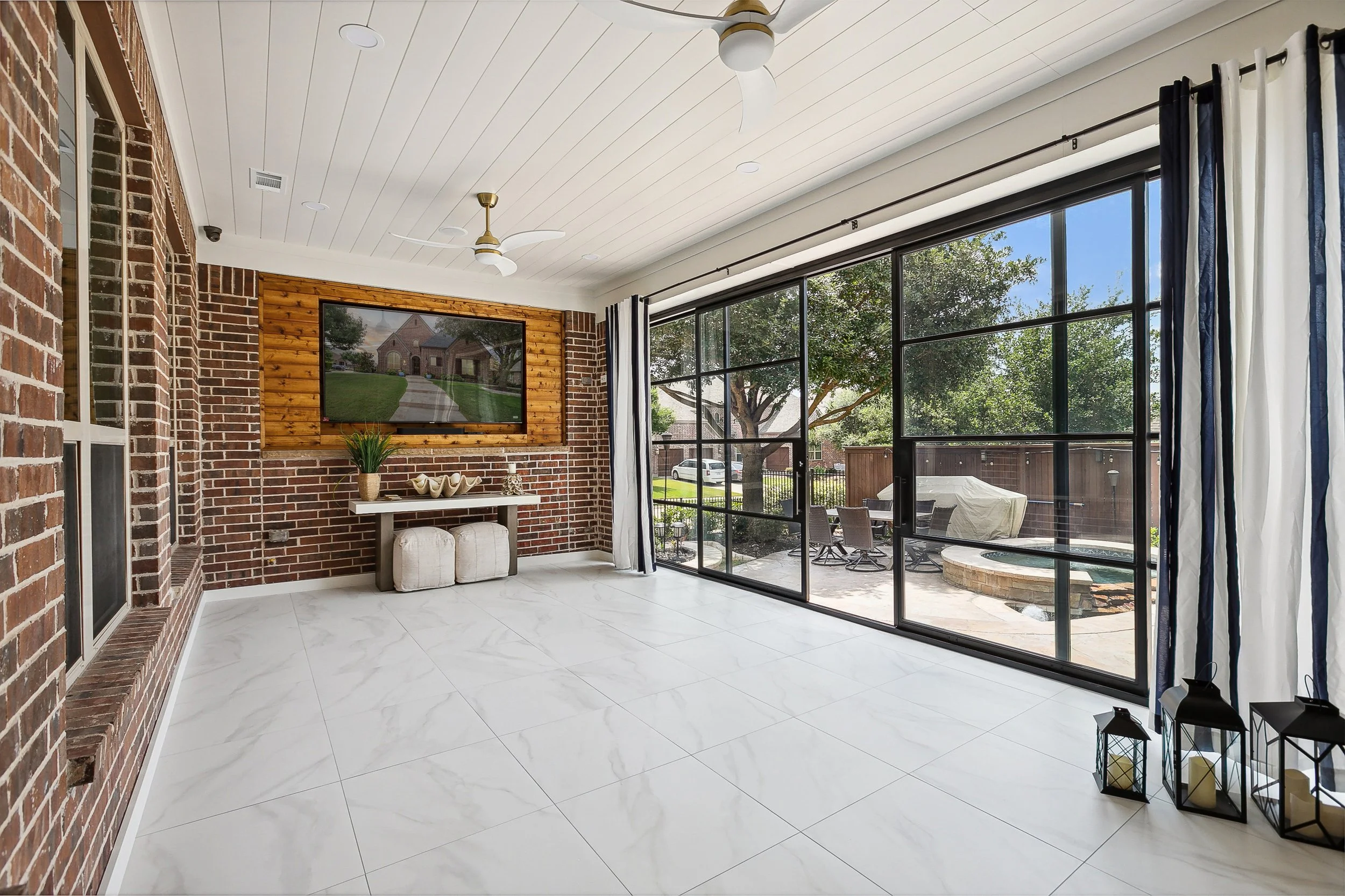 Bright sunroom with large windows, brick walls, white ceiling fan, mounted flat-screen TV on wooden wall panel, white table with decorative objects, and outdoor patio with table and chairs visible through glass doors.