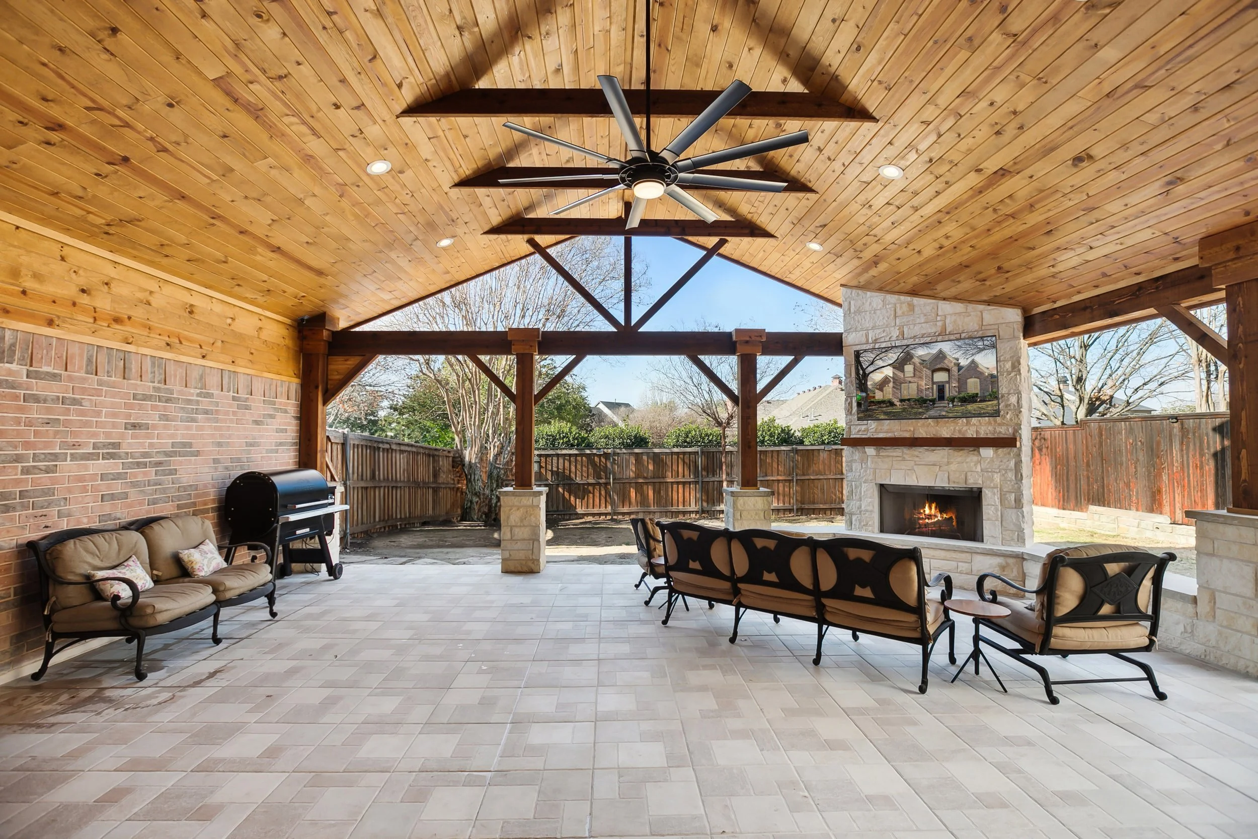 Covered outdoor patio with a wooden ceiling, ceiling fan, brick and stone fireplace, outdoor furniture, and a view of neighboring houses and trees.