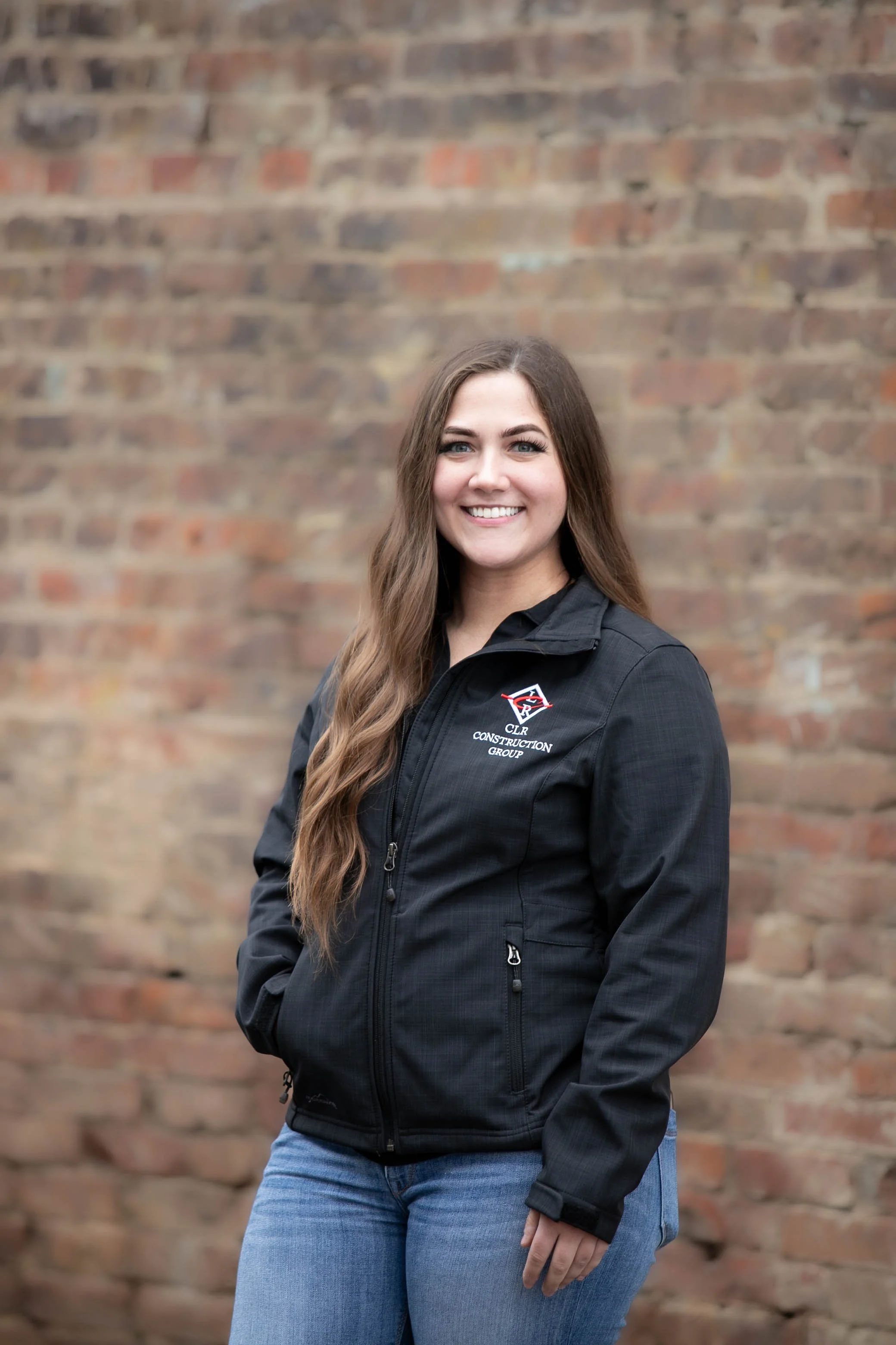 A young woman with long brown hair and blue eyes is smiling and standing in front of a blurry brick wall. She is wearing a black jacket with a logo on the chest and blue jeans.