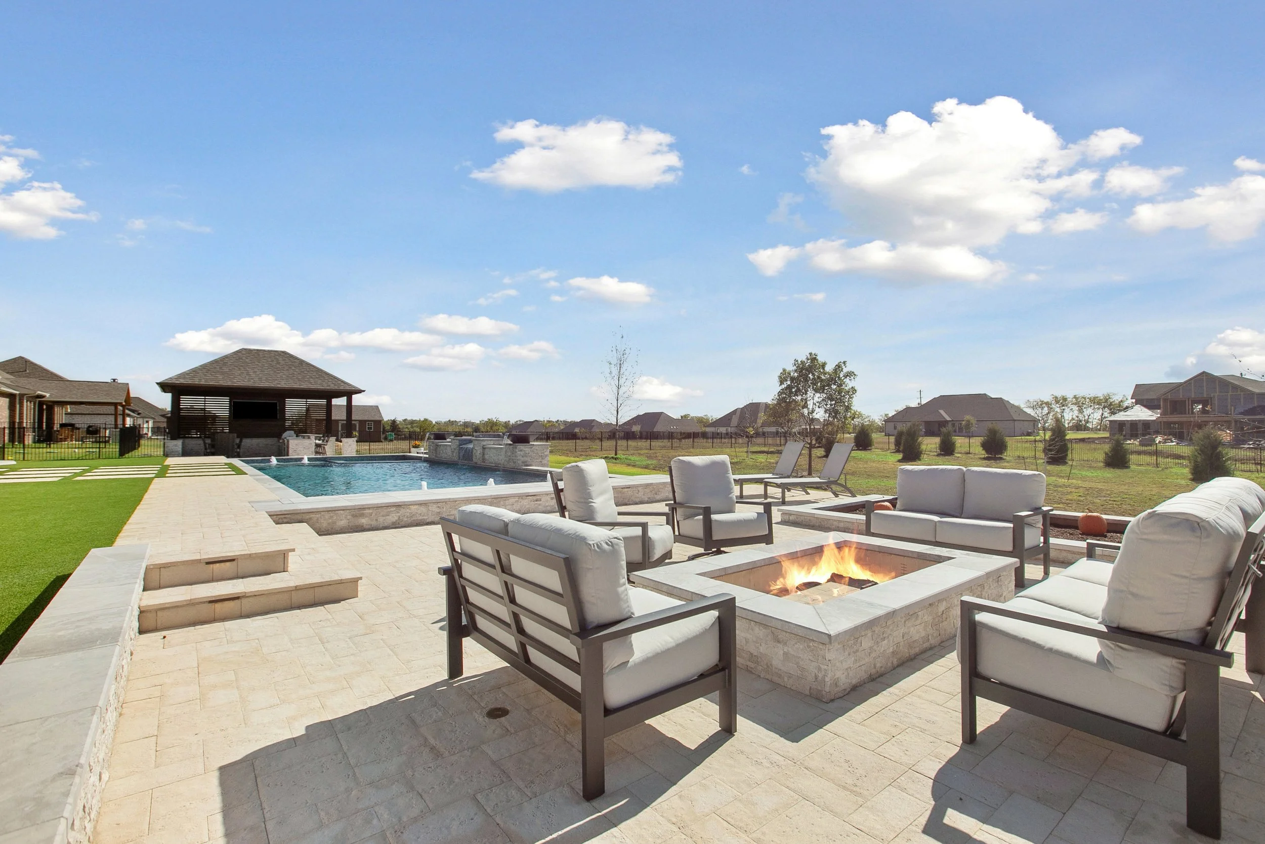Backyard with a swimming pool, outdoor fire pit, patio furniture, and a pavilion under a blue sky with clouds.