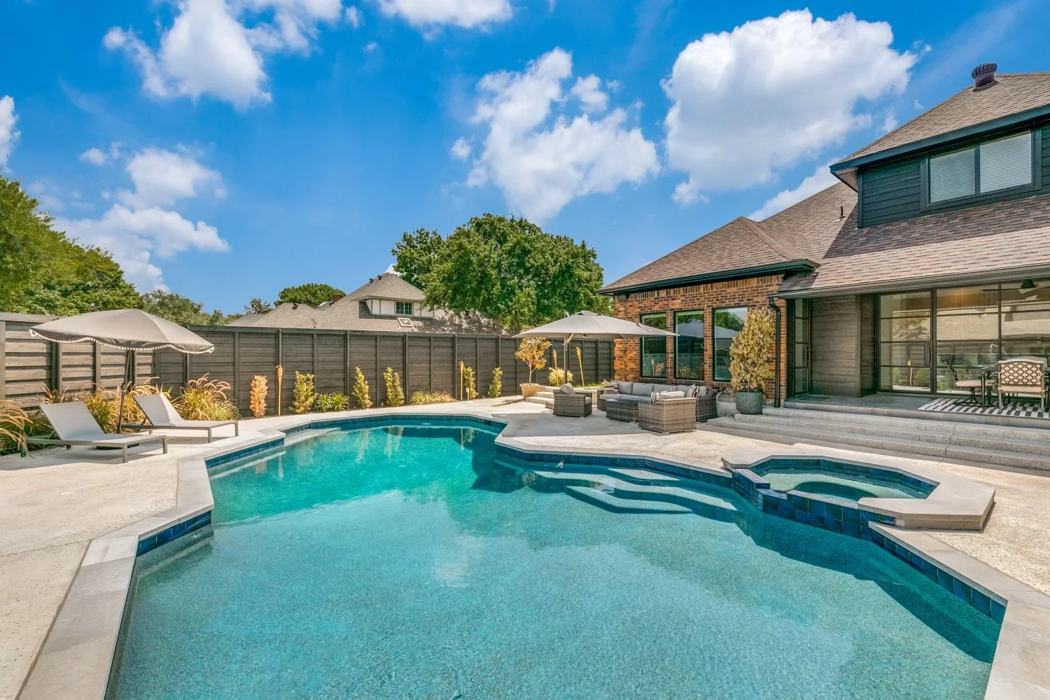 Backyard with swimming pool, patio furniture, and house under blue sky with clouds.