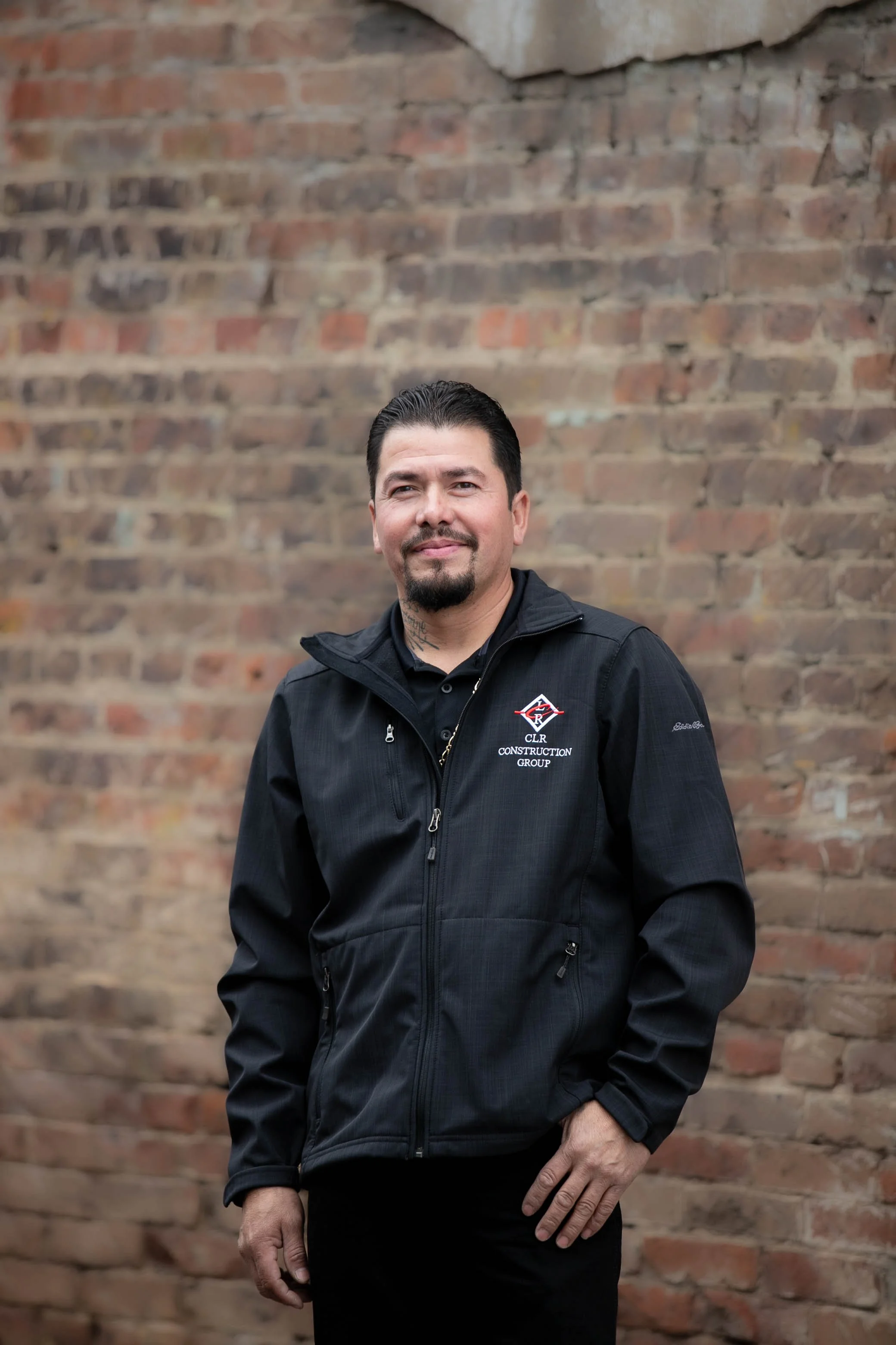 A man wearing a black jacket with company logo and standing in front of an exposed brick wall.
