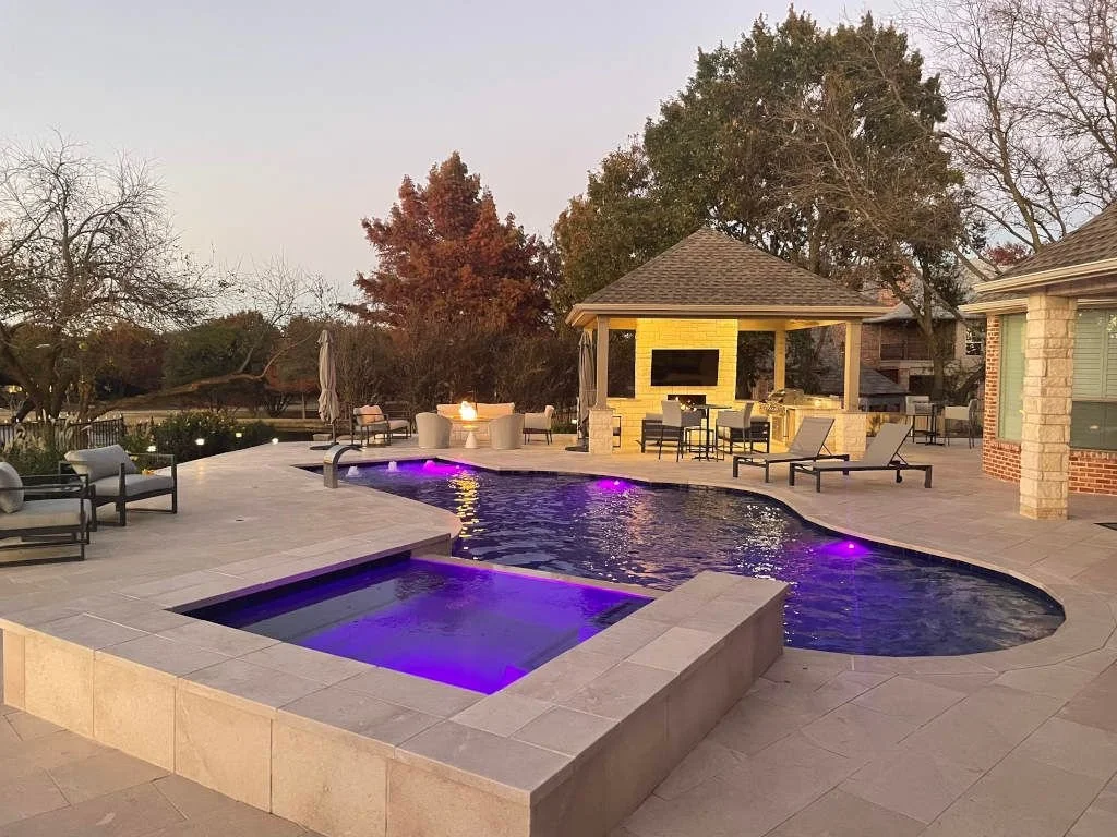 An outdoor pool area with a hot tub, lounge chairs, and a covered bar with a television, surrounded by trees and evening sky.