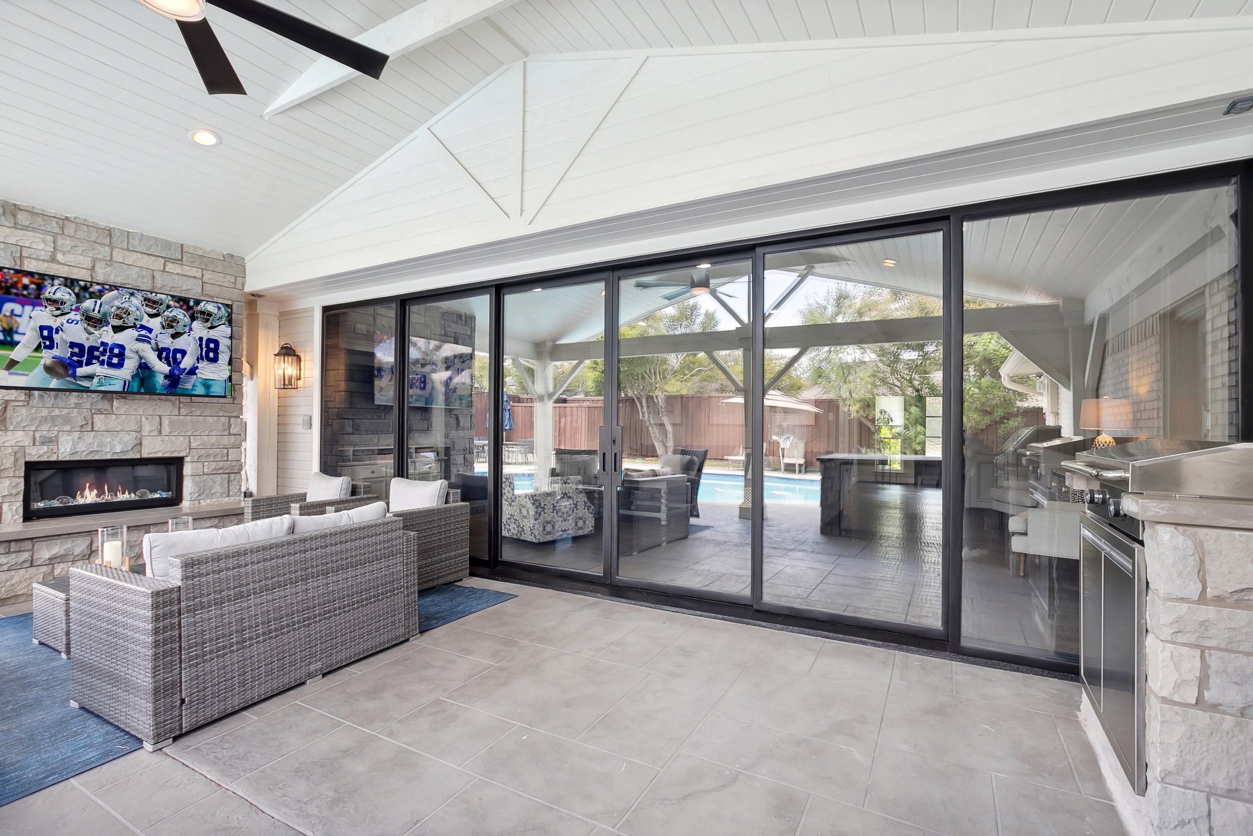 Indoor patio with gray tiled floor, wicker seating, a stone fireplace, large glass sliding doors leading outside, and a ceiling fan.