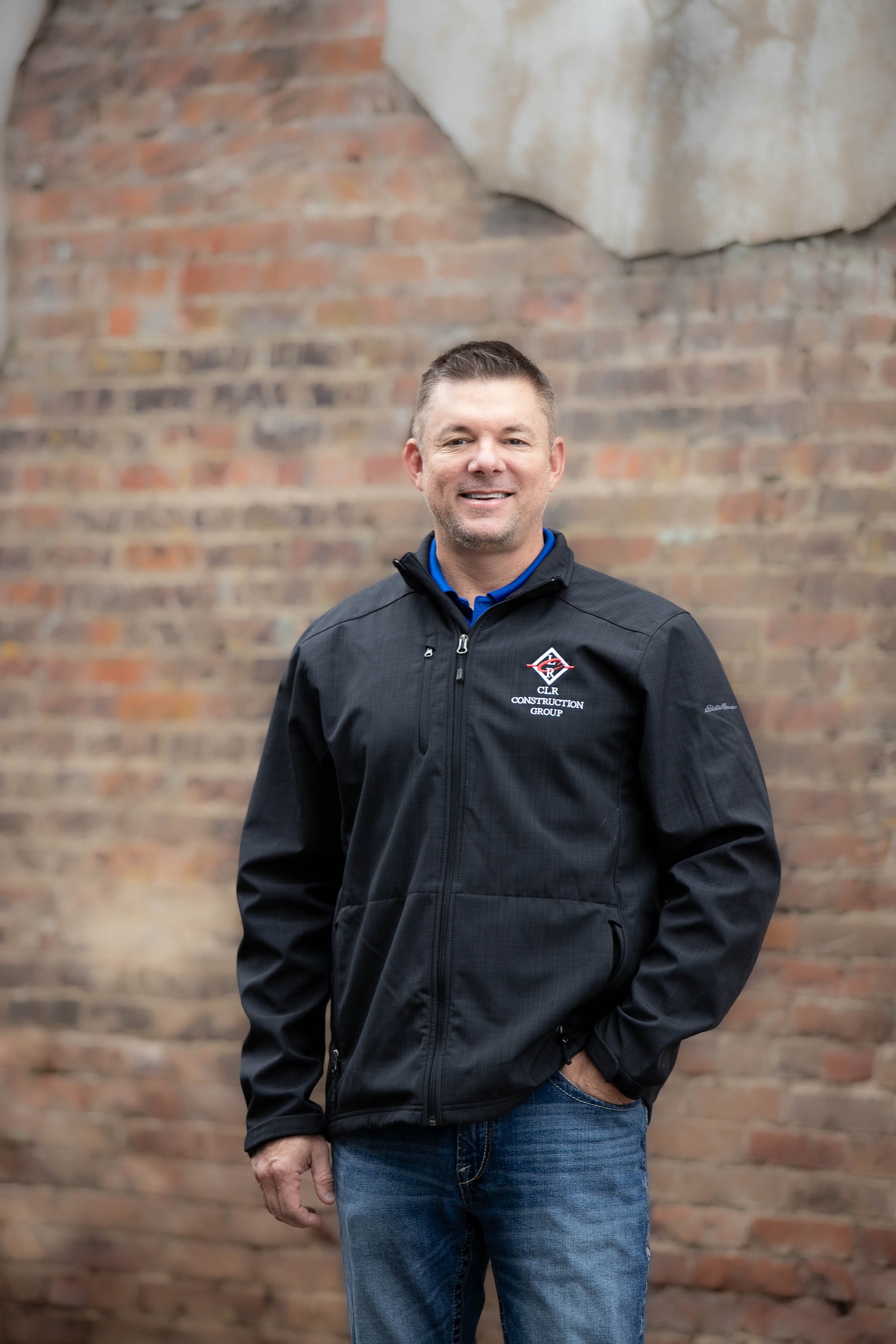A man wearing a black jacket with a construction group logo, standing in front of a brick wall, smiling at the camera.