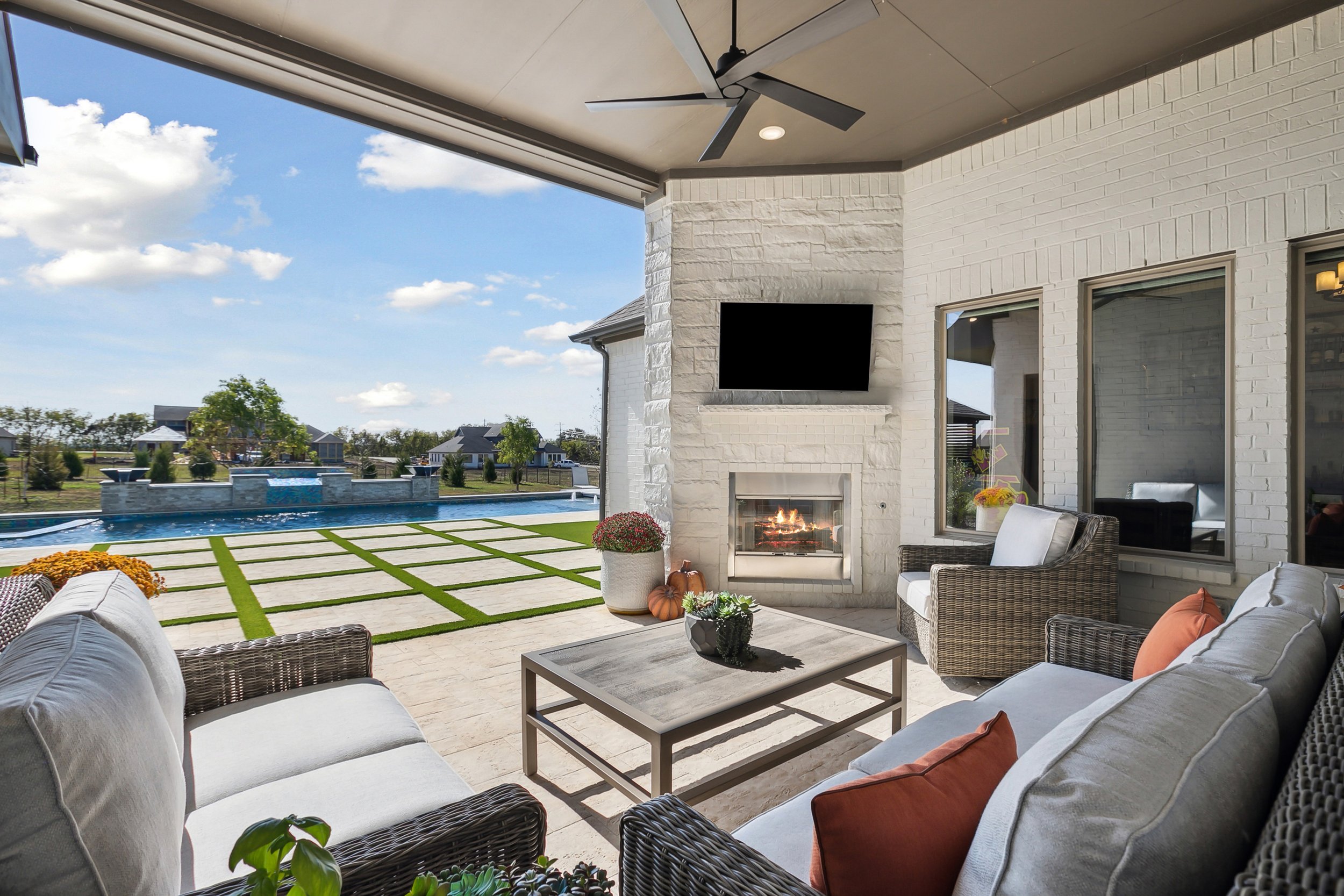 Outdoor patio with wicker furniture, a television mounted on a brick chimney, a fireplace, and a ceiling fan, overlooking a pool and a backyard with houses and trees under a blue sky with clouds.